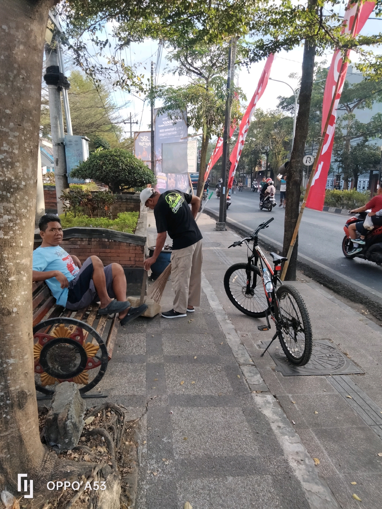 In the city of Kelutan, Indonesia, a man in a light blue t-shirt and dark shorts is sitting on a wooden bench, looking forward. Beside him, a person wearing a white cap and a black t-shirt with a neon green graphic is sweeping the sidewalk with a broom. A bicycle with a water bottle attached to its frame is parked upright on the sidewalk next to the sweeping man. In the background, a street with several motorcycles and some pedestrians is visible, lined with red and white flags hanging from poles. The scene is set outdoors with trees providing shade and a tiled walkway.