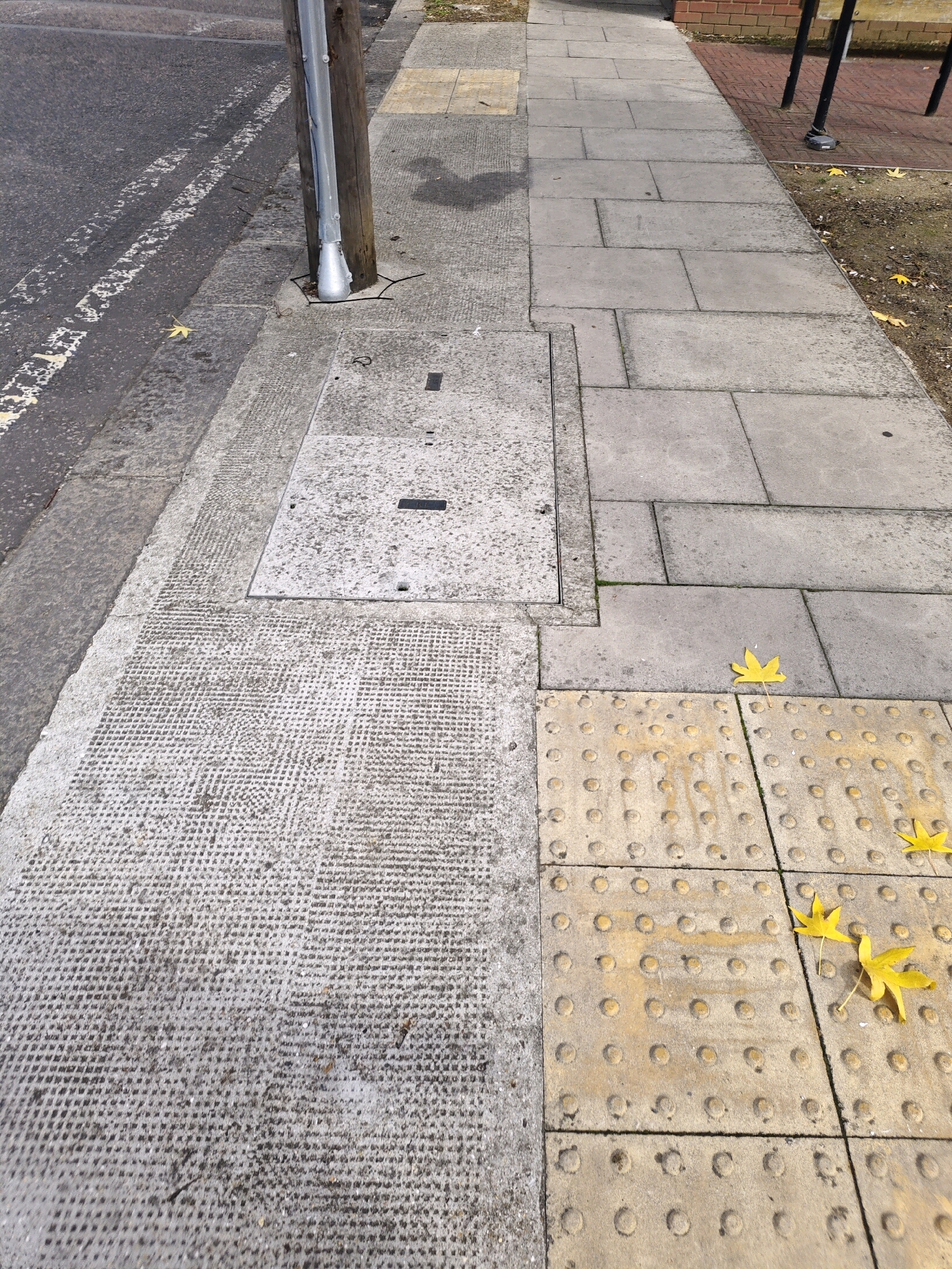 The image shows a section of a pavement on a street. The pavement is made of concrete slabs, with tactile paving indicators on the right side. Several fallen yellow leaves are scattered on the pavement. A manhole cover is visible on the pavement, as well as a metal pole with a conduit attached to it. The street is to the left of the pavement, with white lines marking the traffic lanes. In the background, there is a brick wall and some railings. The scene appears to be in an urban environment, possibly a residential street or a public walkway. The lighting suggests it is daytime, with sunlight casting shadows. There are no people visible in the image. The location is not specified in the image.