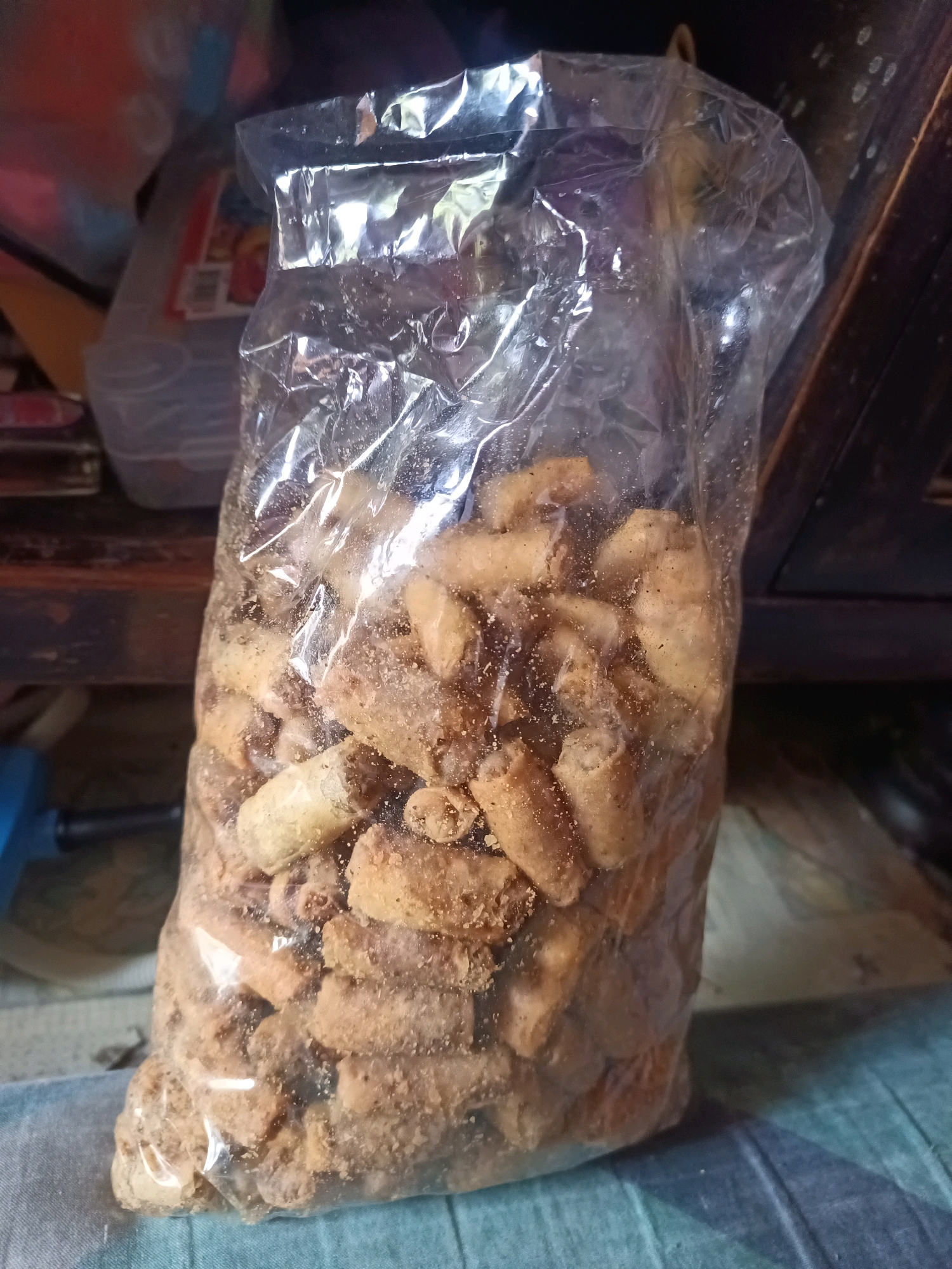 A clear plastic bag filled with numerous small, irregularly shaped, light brown fried snacks is positioned centrally in the frame. The snacks appear to be coated in a fine powder or seasoning. The bag is sealed at the top and has a slightly crinkled texture. In the background, out of focus, are various items including what appears to be another plastic container with red and white labeling, and dark wooden furniture or shelving. The scene is dimly lit, suggesting an indoor environment. The bag of snacks is resting on a blue and green patterned surface. The overall impression is that of a casual, domestic setting, likely a kitchen or pantry area. The image does not provide any specific visual cues that would indicate its location within South Jakarta, Indonesia.