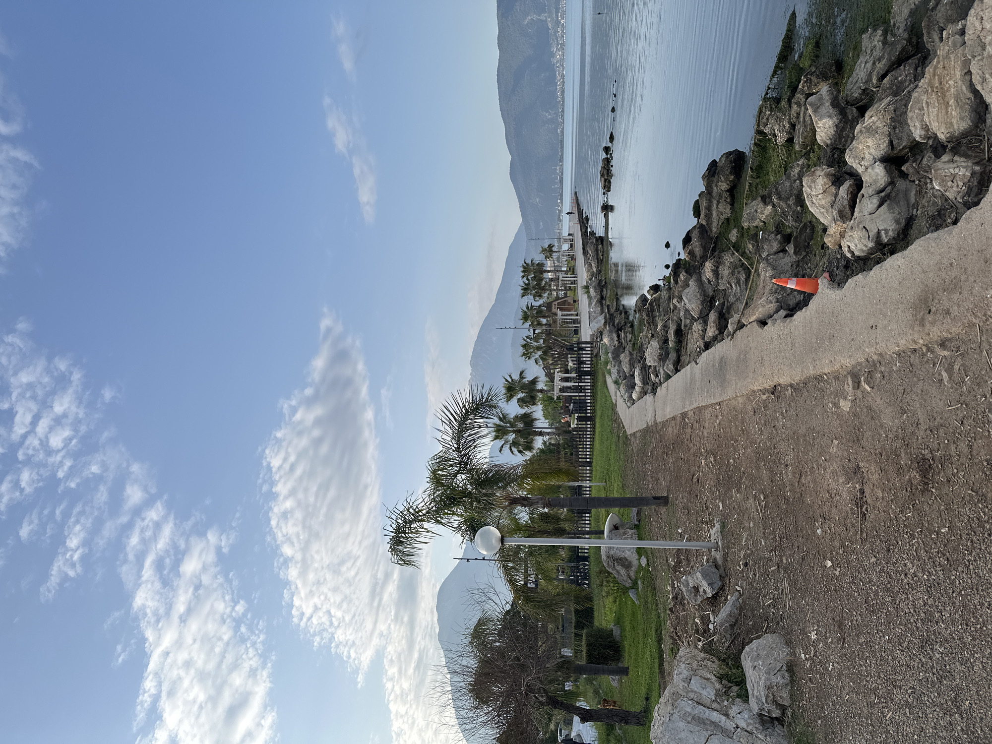 The image depicts a waterfront area in Fethiye, Turkey, with a view of the sea, mountains, and sky. A concrete path runs alongside a rocky barrier separating the path from the water. An orange traffic cone sits on the rocks. Several palm trees and streetlights are visible along the path. The sky is partly cloudy. The mountains are in the background. The foreground consists of dirt and grass.
