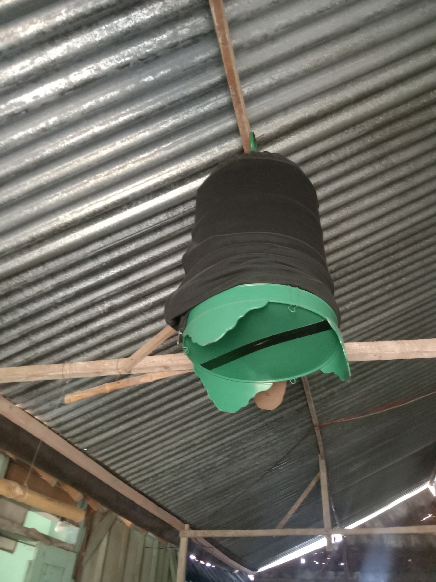 A black fabric funnel is attached to a green plastic bucket, suspended from a corrugated metal roof by wooden beams. The scene is indoors, likely a workshop or storage area, with visible wooden structures and lighting fixtures. The overall impression is of a makeshift or utilitarian setup.