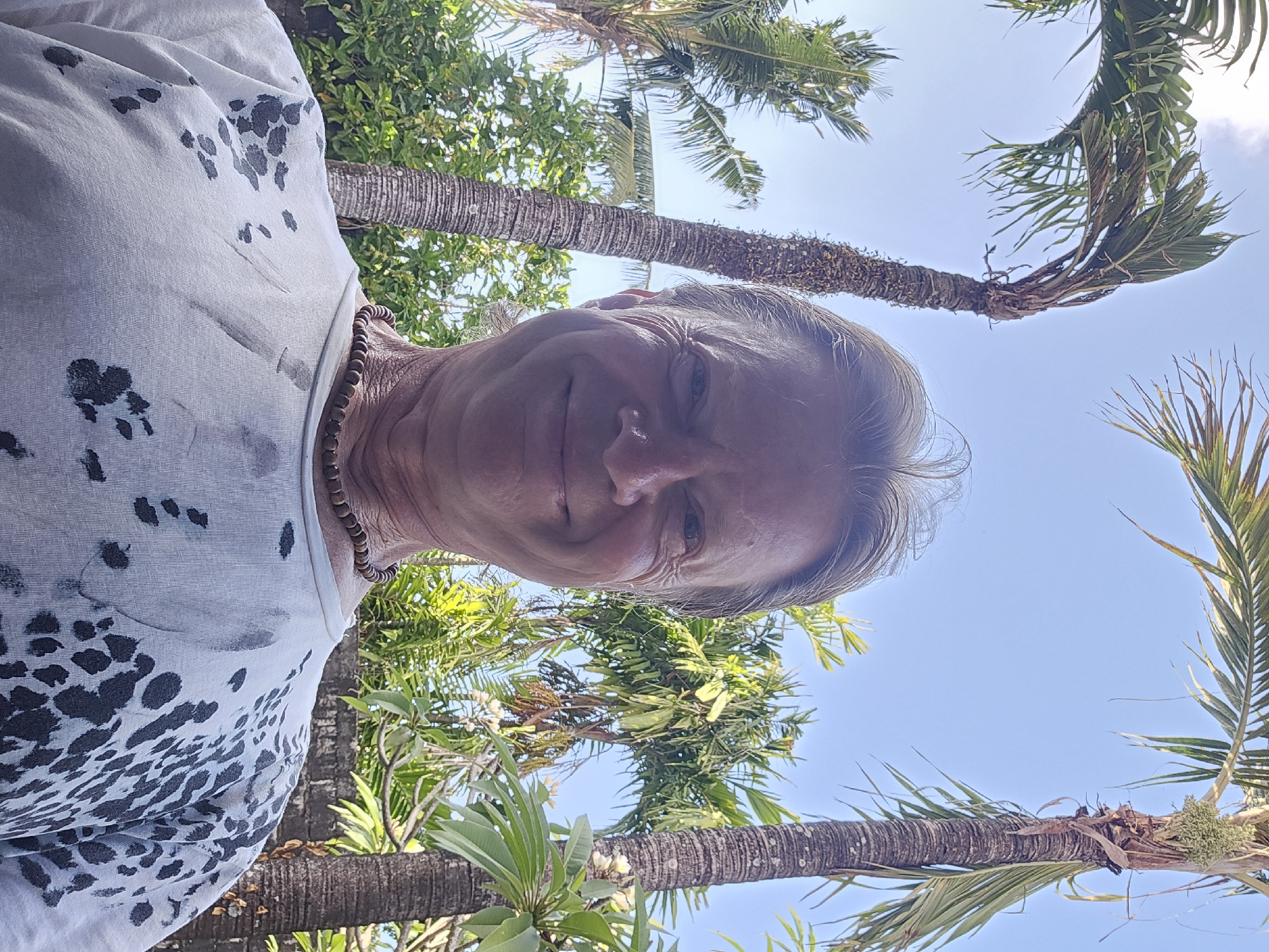 A man with blond hair and a beaded necklace smiles at the camera, wearing a white t-shirt with a black, splotchy pattern. He stands outdoors, framed by palm trees and lush greenery under a clear blue sky. The scene appears to be in Denpasar, Indonesia, suggested by the presence of tropical vegetation and a brick wall in the background.