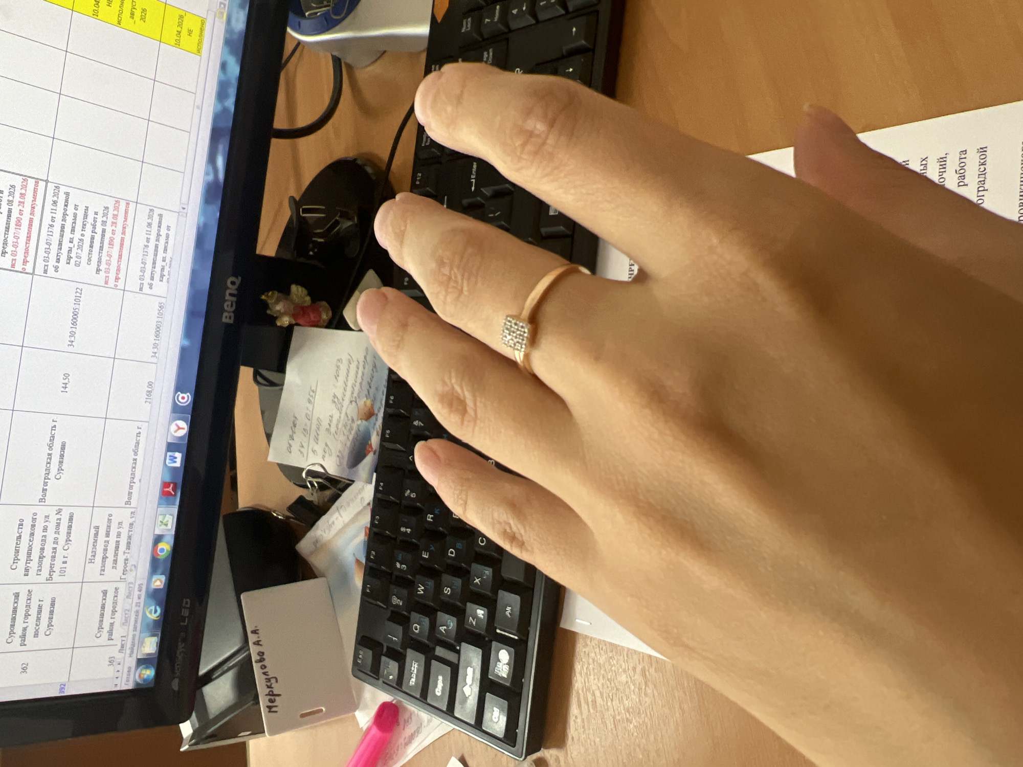 The image shows a person's right hand wearing a ring with a small square diamond, resting on a keyboard. The background includes a computer monitor displaying a spreadsheet or table with Cyrillic text, likely from Volgograd, Russia, as evidenced by the text "Волгоградская область" (Volgograd Oblast). There are also office supplies such as a white card with Cyrillic text that reads "Меркулова А.А." (Merkulova A.A.), a pink pen, and other papers. The scene appears to be a desk in an office environment. The computer screen shows entries related to construction, gas pipelines, and road construction, with dates and document numbers.