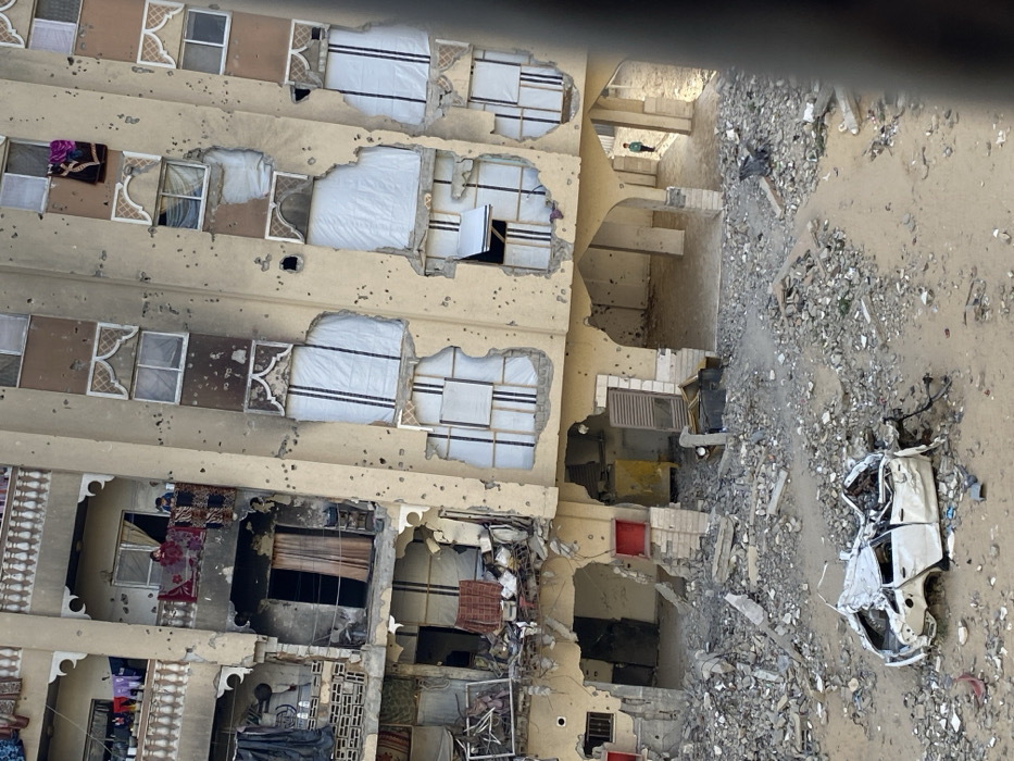 An outdoor, high-angle view depicts a severely damaged multi-story residential building located in Khan Yunis, Palestinian Territory. The building's beige-toned facade is heavily pockmarked with countless impacts consistent with bullets or shrapnel. Multiple sections of the building's outer walls are completely blown away, exposing interior rooms with rubble, damaged fixtures, and personal belongings. Balconies on various levels show significant structural damage and debris, with laundry hanging on upper railings, including a visible red garment. On a damaged second-floor balcony, a woman in a dark red garment stands alongside a smaller figure, both looking out towards the right. Numerous windows are either shattered, missing, or covered with white, opaque material, some framed by decorative, arched patterns that are also damaged.
The ground in front of the building is covered in a dense layer of sand, dirt, and fragmented concrete rubble. Amidst this debris, the heavily mangled and crushed frame of a white four-door sedan is prominently visible in the lower center, its roof and body severely collapsed. Further into the building's ground floor, under an arched opening, a small child in a light green top is seen walking away from the building towards the right side of the frame. The entire scene indicates extensive structural destruction and environmental disruption.