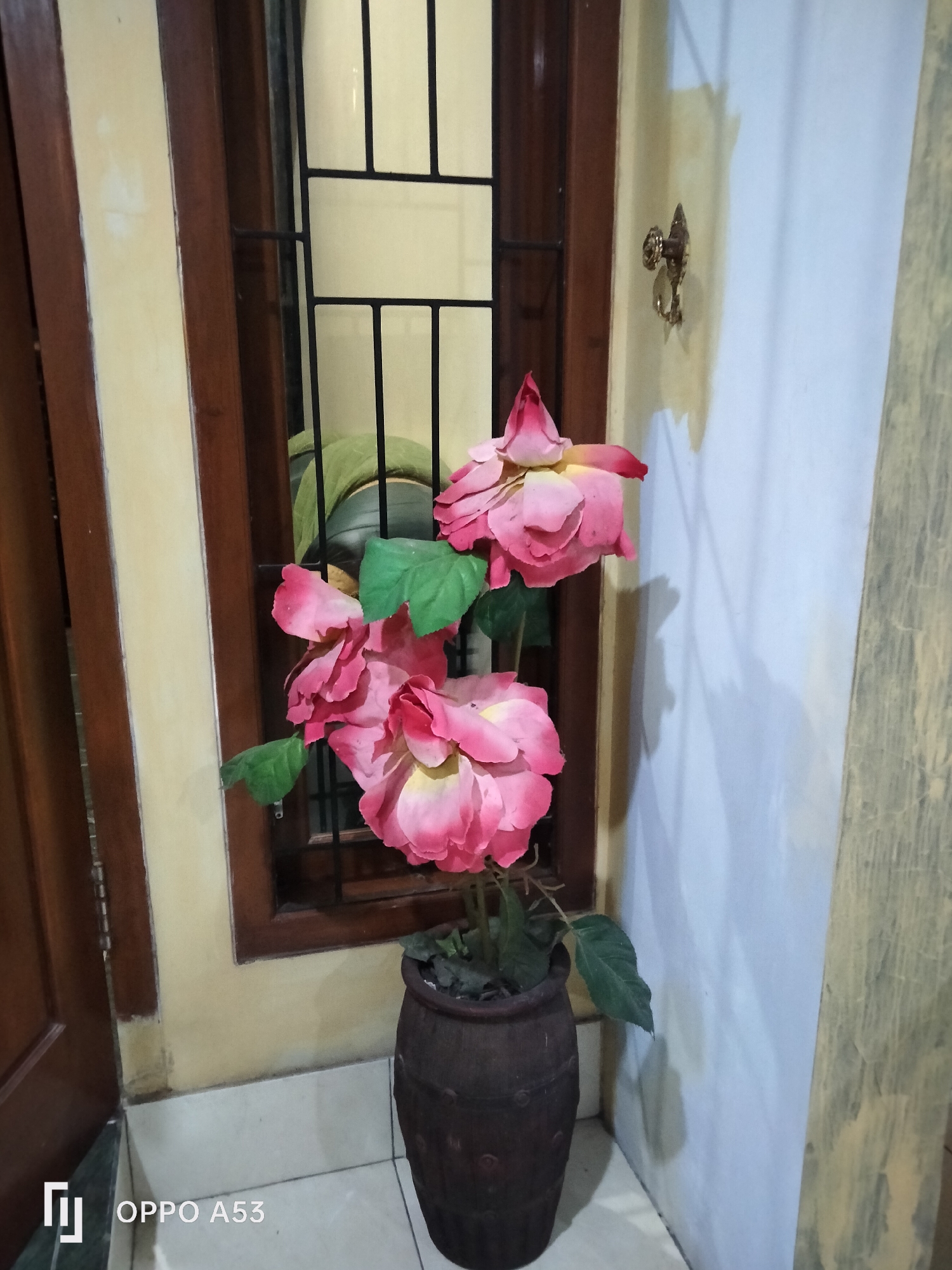 A decorative potted plant with artificial pink roses stands in a dimly lit interior space. The plant is in a dark brown, textured pot. The backdrop includes a wooden-framed window with black bars and a textured wall. To the left, a dark wooden door is partially open, revealing a glimpse of the outside. A decorative metal hook is mounted on the wall to the right of the window. The floor is tiled in a light color. The overall impression is of a domestic setting, potentially in a home or establishment in Rejowinangun, Indonesia, as indicated by the watermark at the bottom left of the image.