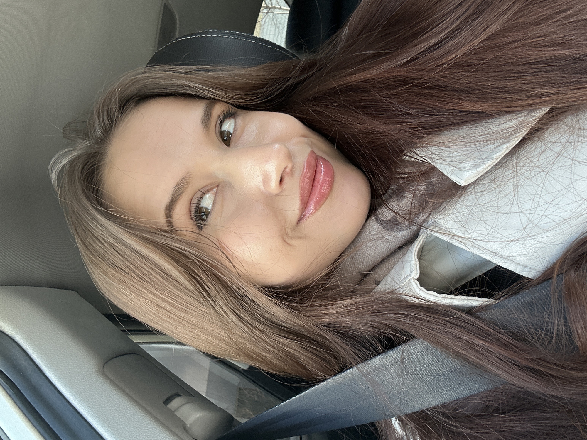 A young woman is captured in a close-up selfie or portrait taken inside a car in Yekaterinburg, Russia. Her head is tilted, giving the impression she is lying down or leaning back, though she is seated upright with a seatbelt visible across her chest. She possesses long, wavy dark brown hair, with some lighter strands framing her face. She looks slightly to her right (viewer's left) with a gentle, inviting smile that subtly reveals her teeth. Her makeup includes glossy pinkish-nude lipstick, mascara, and defined eyebrows.
She is dressed in a light-colored, possibly white or off-white, collared shirt or jacket, layered over a grey turtleneck or scarf. A grey seatbelt is prominent across her body. The car's interior is partially visible, showing a black leather headrest with white stitching behind her, and grey plastic trim on the car's door panel and A-pillar. Natural light illuminates her face, indicating it is daytime. The overall expression is pleasant and contented. No text is visible in the image.