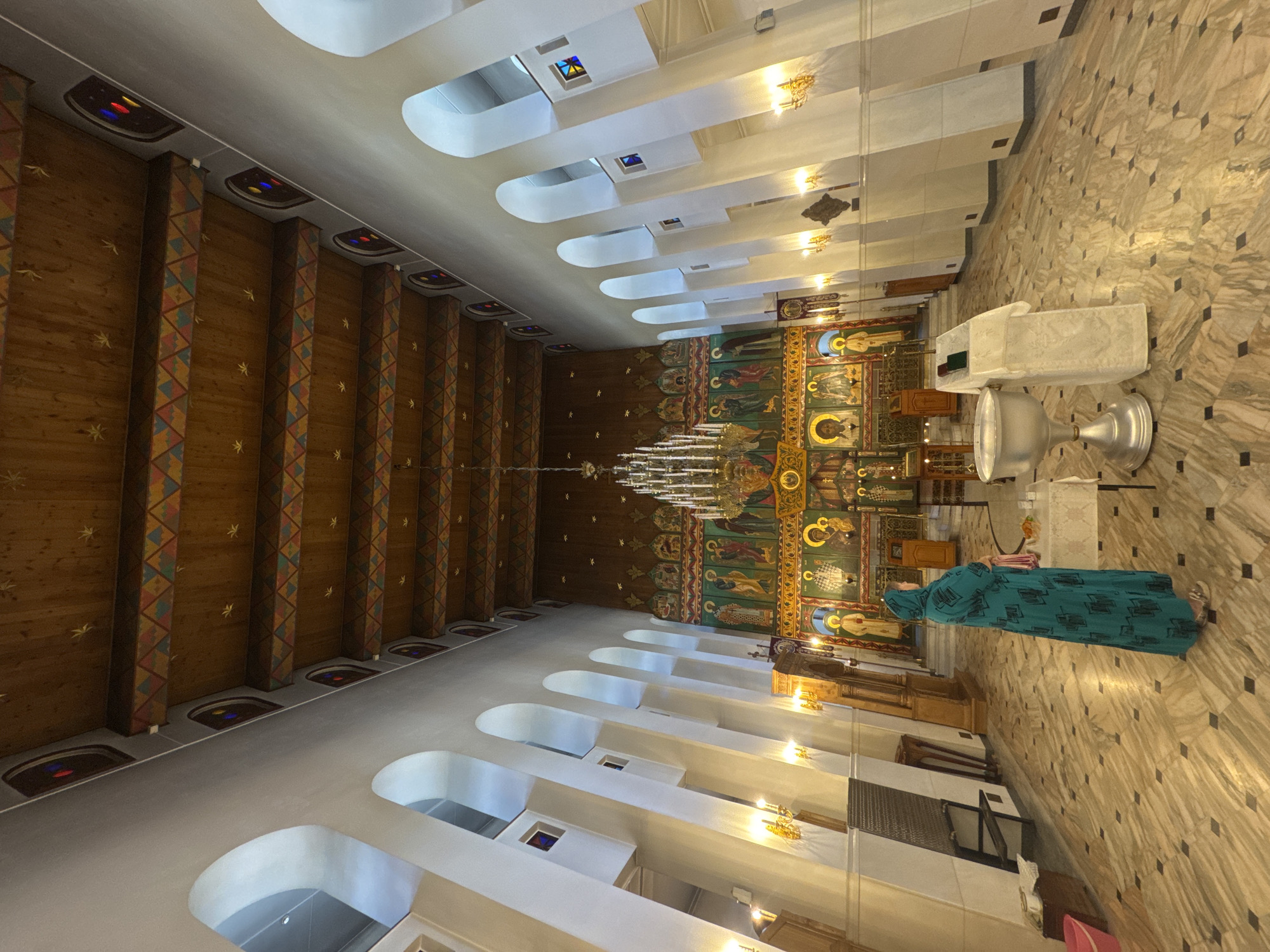 The image shows the interior of the Transfiguration Cathedral in Tolyatti, Russia. A woman wearing a long, teal dress with a headscarf stands facing the altar. In the foreground, a baptismal font is visible, along with a table covered with a white cloth. The cathedral features a high wooden ceiling with decorative beams and stained-glass windows. The walls are adorned with religious iconography. The floor is made of marble with a checkered pattern. The overall impression is one of solemnity and spiritual grandeur.