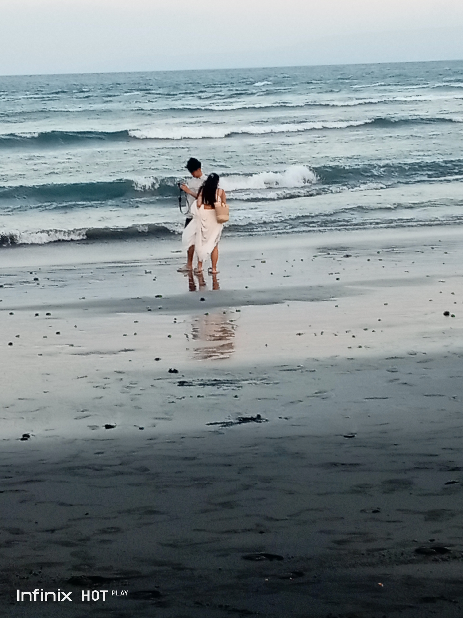 In Blahbatuh, Indonesia, at the beach, a man and a woman stand near the water's edge. The man, facing away from the camera, is wearing a white shirt. The woman, also facing away, is in a white dress and holds a straw bag. They are positioned on wet sand, with gentle waves approaching them. The water is calm with small ripples. The sky above is overcast and a pale gray. The sandy shore is dark and reflective, with scattered small dark objects. The Infinix HOT PLAY logo is visible in the bottom right corner of the image.