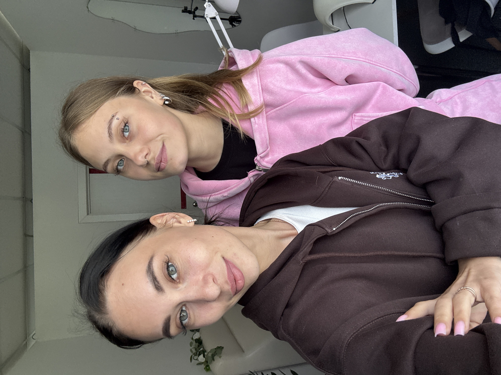 Two young women are posing indoors. The woman on the left has light brown hair, fair skin, and light-colored eyes, with a piercing above her eyebrow and earrings. She is wearing a pink zip-up hoodie over a black top. The woman on the right has dark hair, fair skin, and light-colored eyes, with earrings. She is wearing a brown zip-up hoodie over a white top. They are both looking towards the camera. The background includes a white wall, a plant, and what appears to be a desk or table with various items on it. The location is unspecified, and Фоки, Russia, cannot be confirmed as the location.