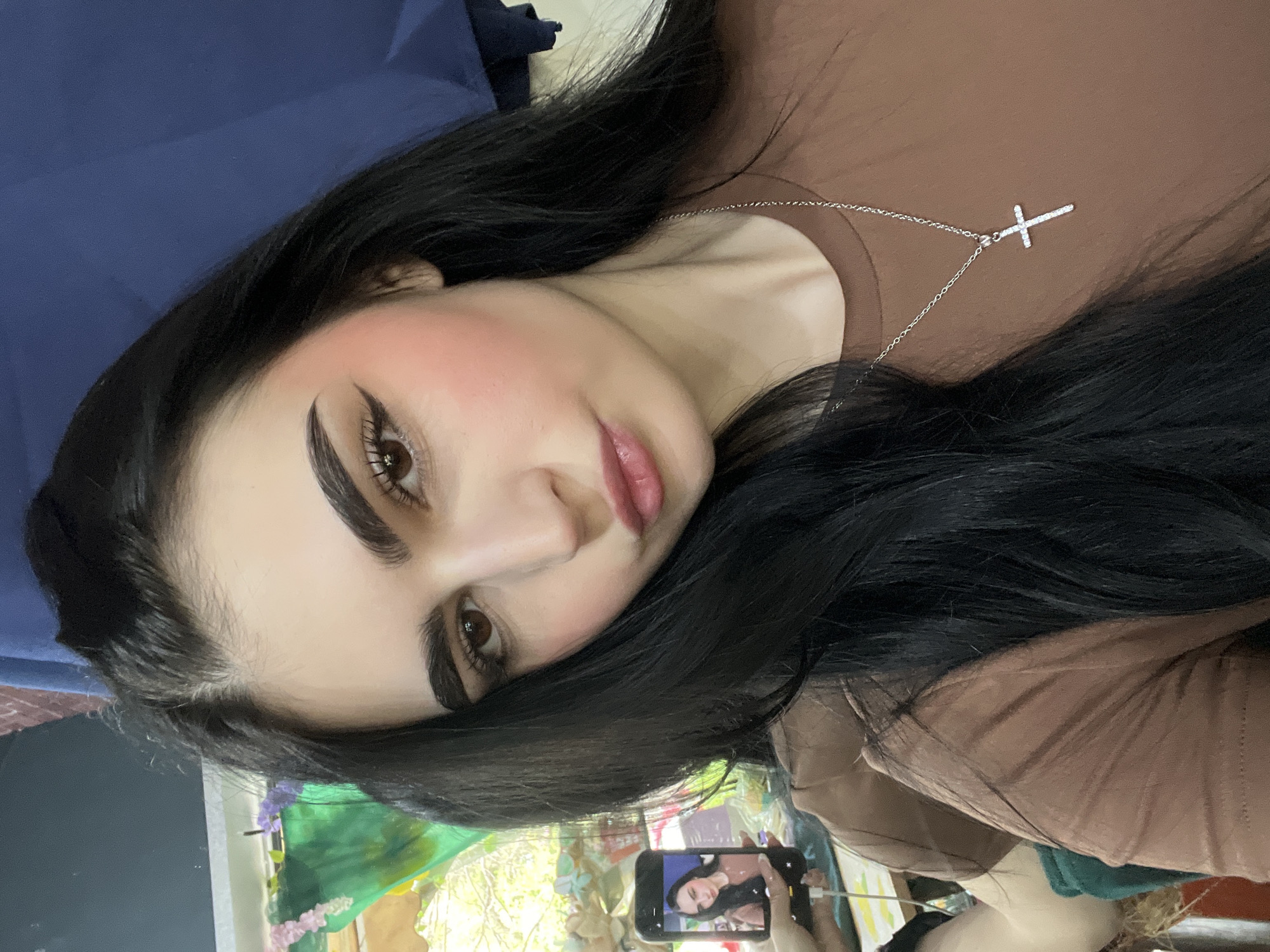 A woman with long, dark hair and fair skin is posing for a selfie. She is wearing a brown top and a silver necklace with a cross pendant. She has dark, defined eyebrows and heavy winged eyeliner. Her cheeks are flushed with pink blush, and her lips are pink. She is holding a smartphone in her hand, which is displaying a smaller version of the same image. The background is blurred, but it appears to be a room with floral curtains and a blue cushion. The location cannot be determined from the image.