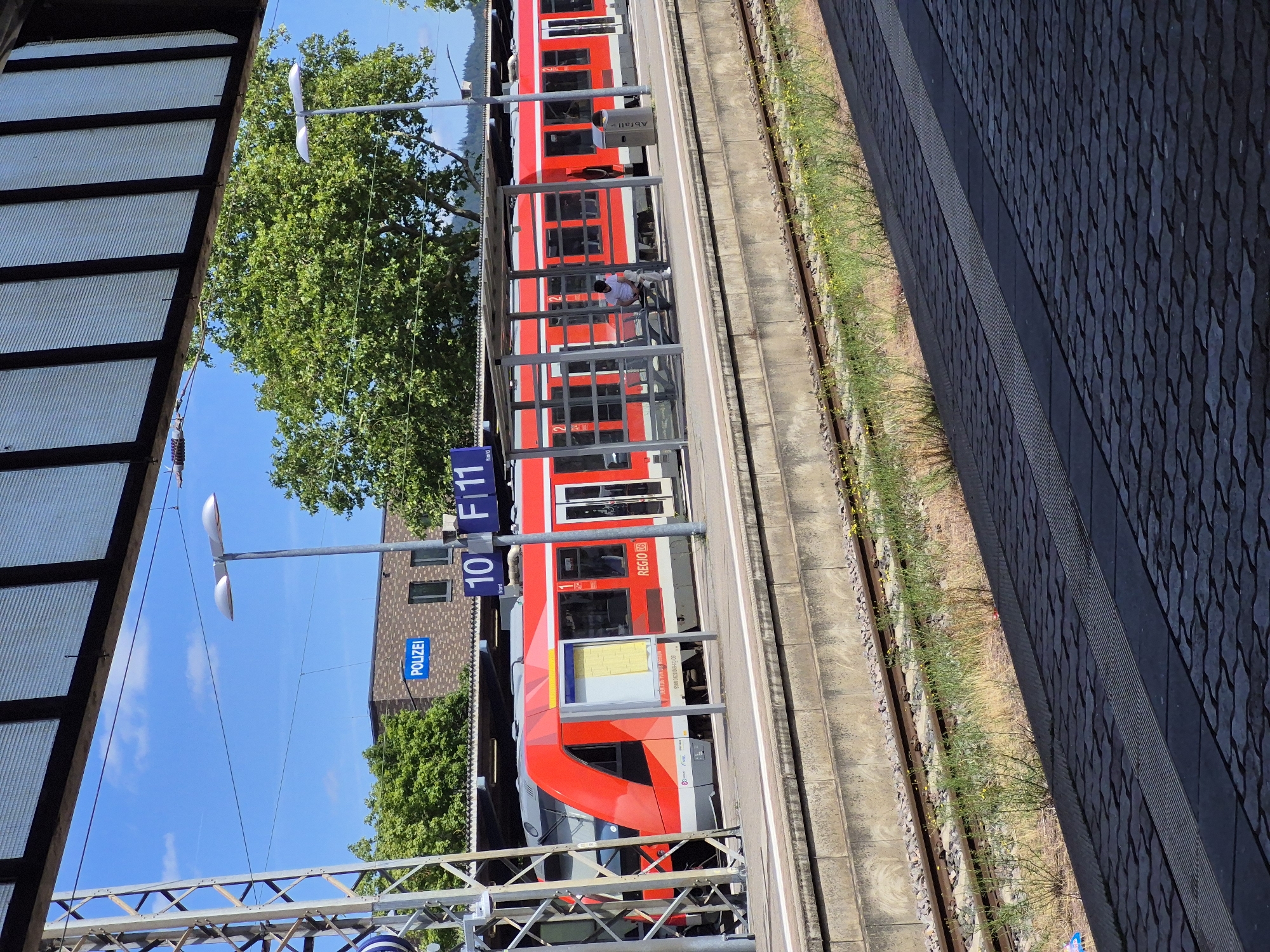 The image shows a red train at a station platform in Trier, Germany. A blue sign with "POLIZEI" indicates a police station or presence nearby. The train is labeled "REGIO DB". There are a few people visible, including one person sitting on a bench under a shelter. The platform is paved with dark tiles, and there are train tracks with gravel and grass. Overhead, a section of a building's roof with translucent panels is visible. The sky is blue with some scattered clouds, and trees are present in the background.