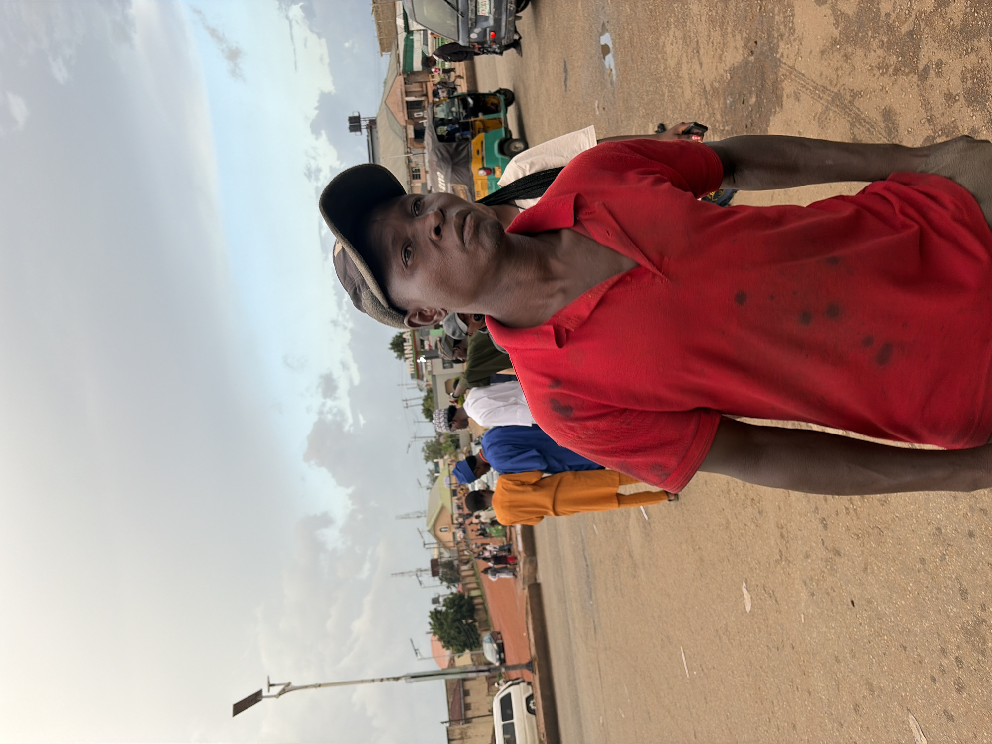 The image depicts a street scene in Gura Top, Nigeria. In the foreground, a man wearing a red polo shirt and a dark baseball cap is the primary subject. He has dark skin and appears to be looking slightly off-camera. The red shirt has several dark, irregular stains. To his left, a queue of people dressed in traditional clothing, including an orange and a blue garment, are visible. In the background, there are buildings, vehicles, and a street light. A colorful auto-rickshaw is also present in the middle ground, alongside other cars. The sky is overcast, suggesting a cloudy or possibly rainy day. The ground is a dusty, unpaved surface, with some puddles visible. The overall impression is of an ordinary day in a bustling Nigerian town.