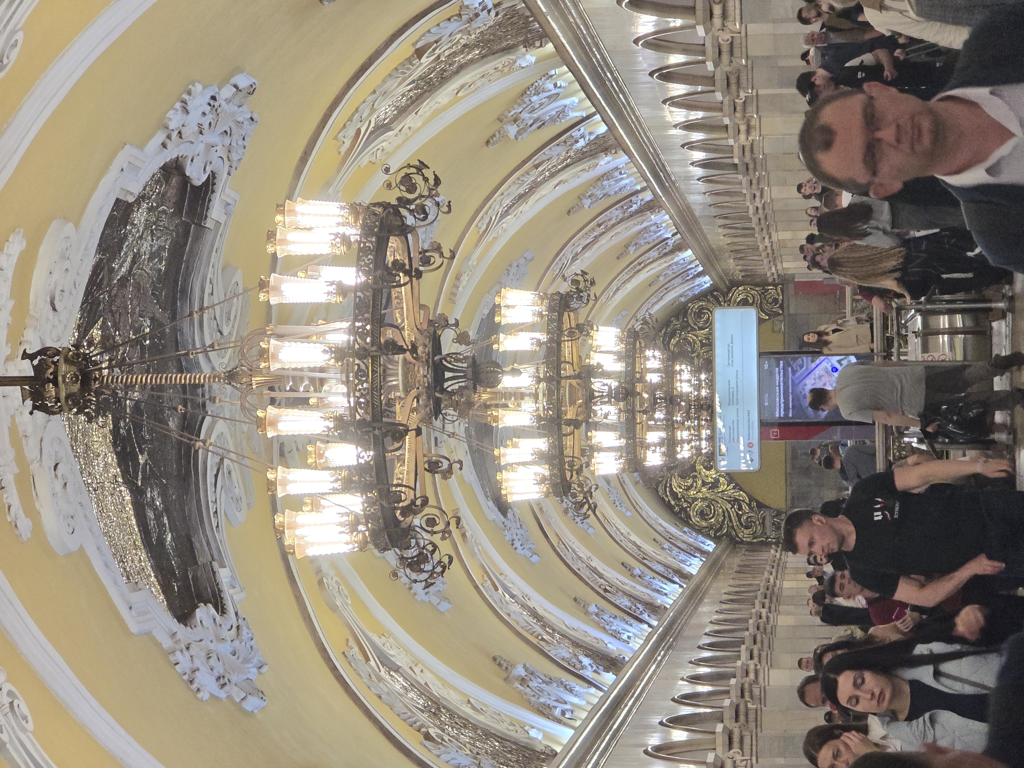 The image depicts a scene within the Moscow Metro, specifically the Komsomolskaya station. The focus is on the station's ornate architectural details, including the high vaulted ceiling adorned with decorative moldings, chandeliers, and ornate embellishments. A significant portion of the image captures a series of large, multi-tiered chandeliers. The ceiling is a pale yellow color, with white and gold accents.
People are present, standing along the platform, some appearing to be waiting for a train, others in transit. There is an electronic screen displaying information, likely train schedules or other relevant announcements. The platform is lined with a balustrade-like structure, possibly serving as a barrier between the platform and the tracks. The overall environment suggests a crowded public space, typical of a metro station.