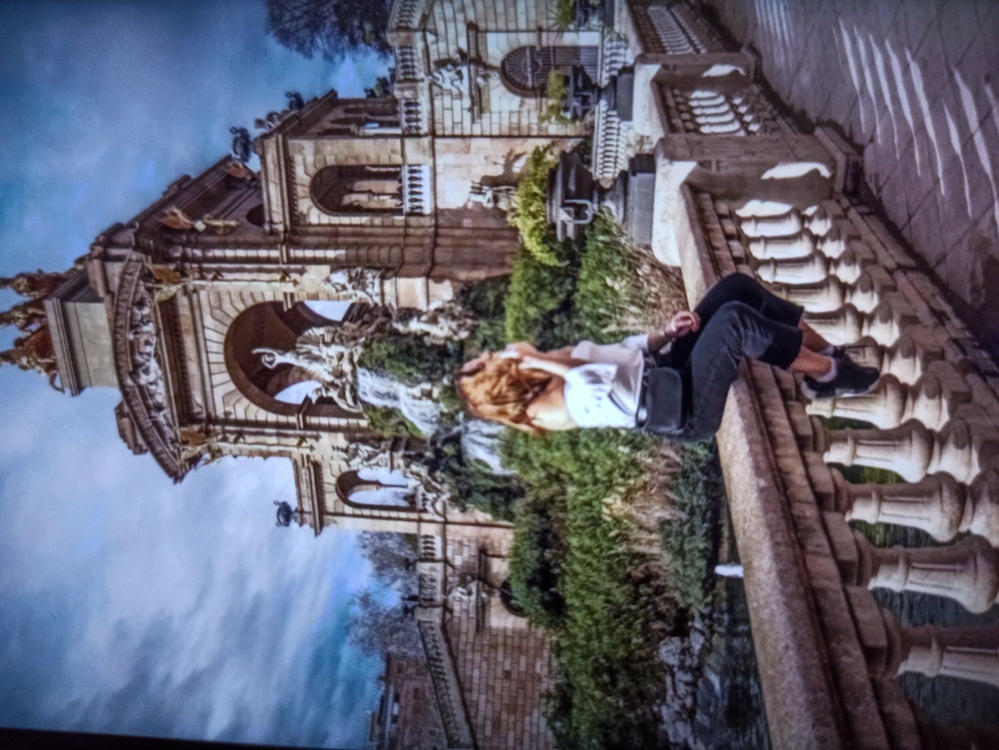 The image depicts a young woman sitting on a stone balustrade with a fountain in the background. The woman is wearing a white off-the-shoulder top and dark pants, with a black fanny pack around her waist. She is looking to her right and holding her hand up to her face. The fountain is elaborate, with multiple tiers and sculptures, surrounded by lush greenery. The sky is cloudy, with dramatic light and shadow patterns cast on the tiled ground. The scene appears to be in a park or public garden. The location is not Balakliya, Ukraine, but rather the Cascada Monumental in Barcelona, Spain.