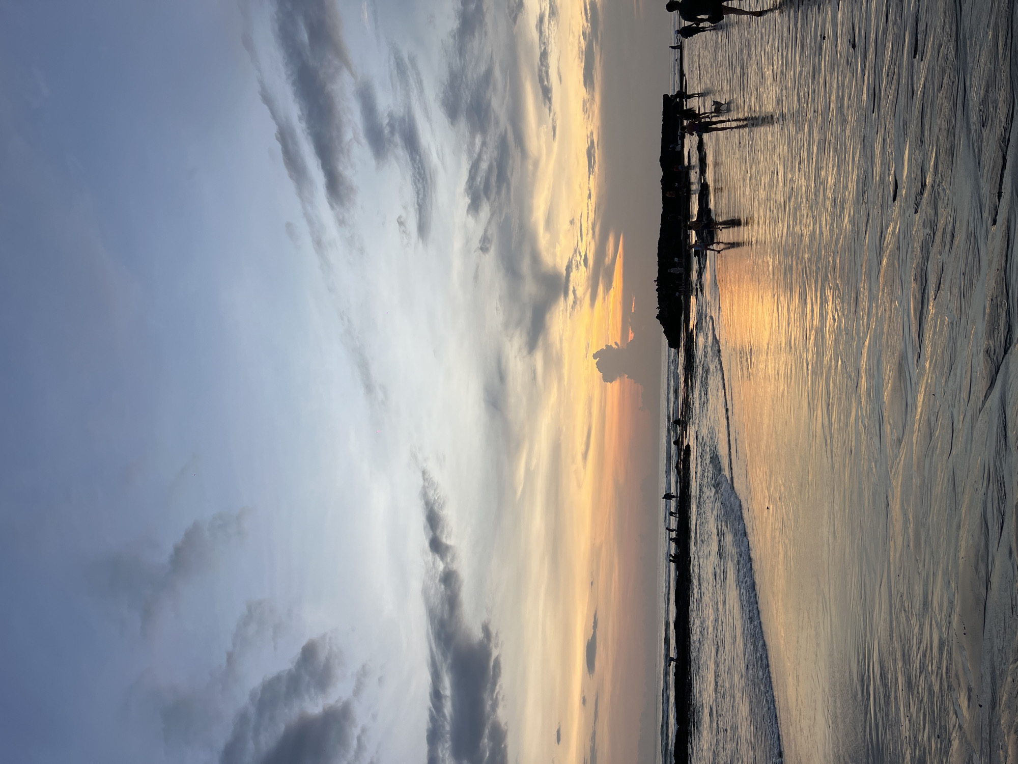 The image shows a beach at sunset in Canggu, Indonesia. The sky is a mix of blue and gray on the left, transitioning to yellow and orange hues near the horizon. Clouds are visible in the sky, some dark gray. Silhouetted figures stand along the shoreline, possibly walking. The wet sand reflects the light from the setting sun. The beach shows patterns created by the receding tide.