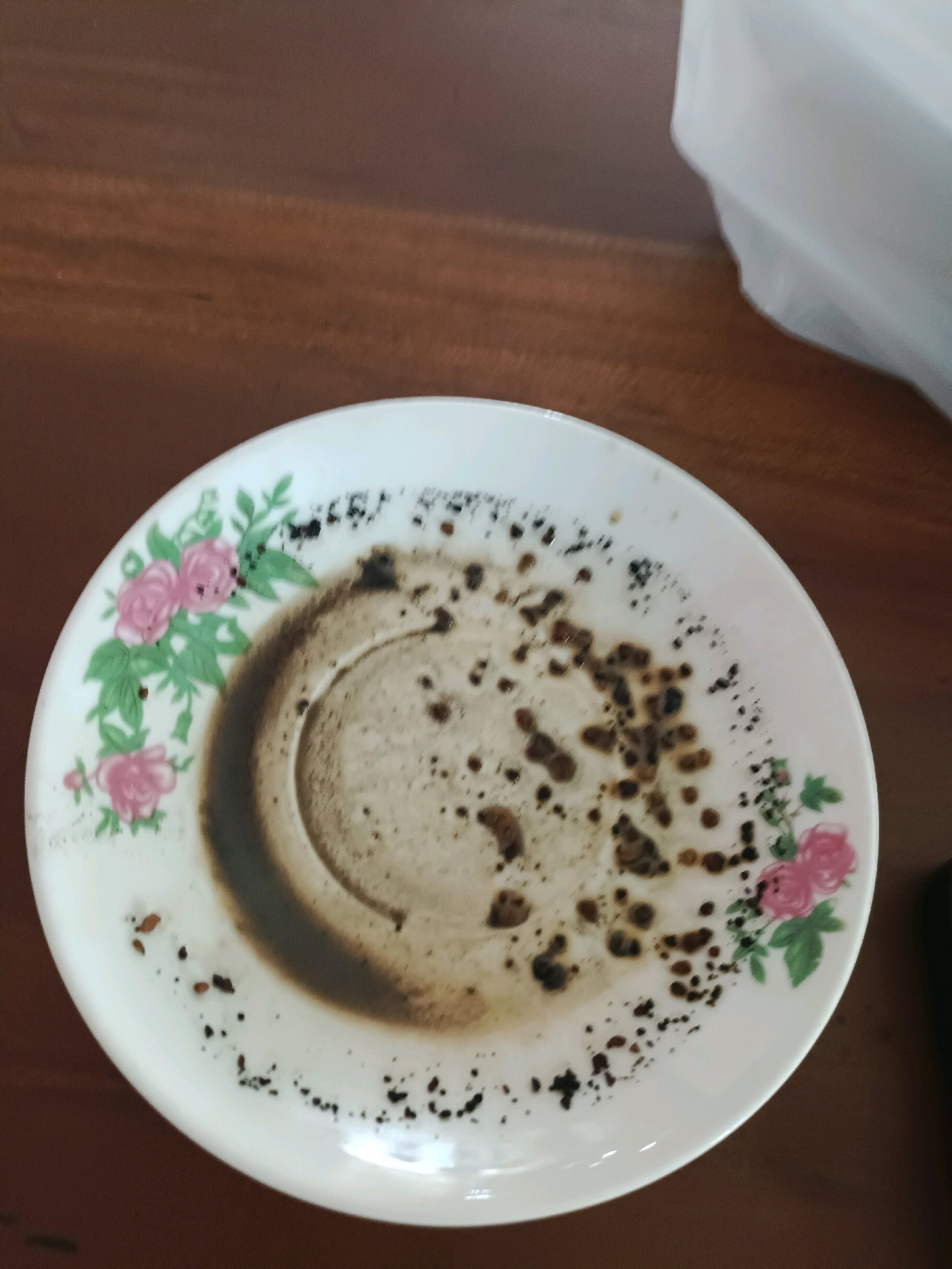 The image shows a close-up, overhead view of a white, ceramic saucer with a floral pattern of pink roses and green leaves. The saucer is resting on a brown, wooden surface. The saucer is dirty, with remnants of dark, granular material scattered across its surface, particularly concentrated in a circular pattern in the center, suggesting it held a beverage like coffee. There are also scattered dark specks and some larger dark clumps on the white surface of the saucer. A blurred white object is partially visible in the upper right corner of the image. The scene appears to be set indoors, possibly on a table.