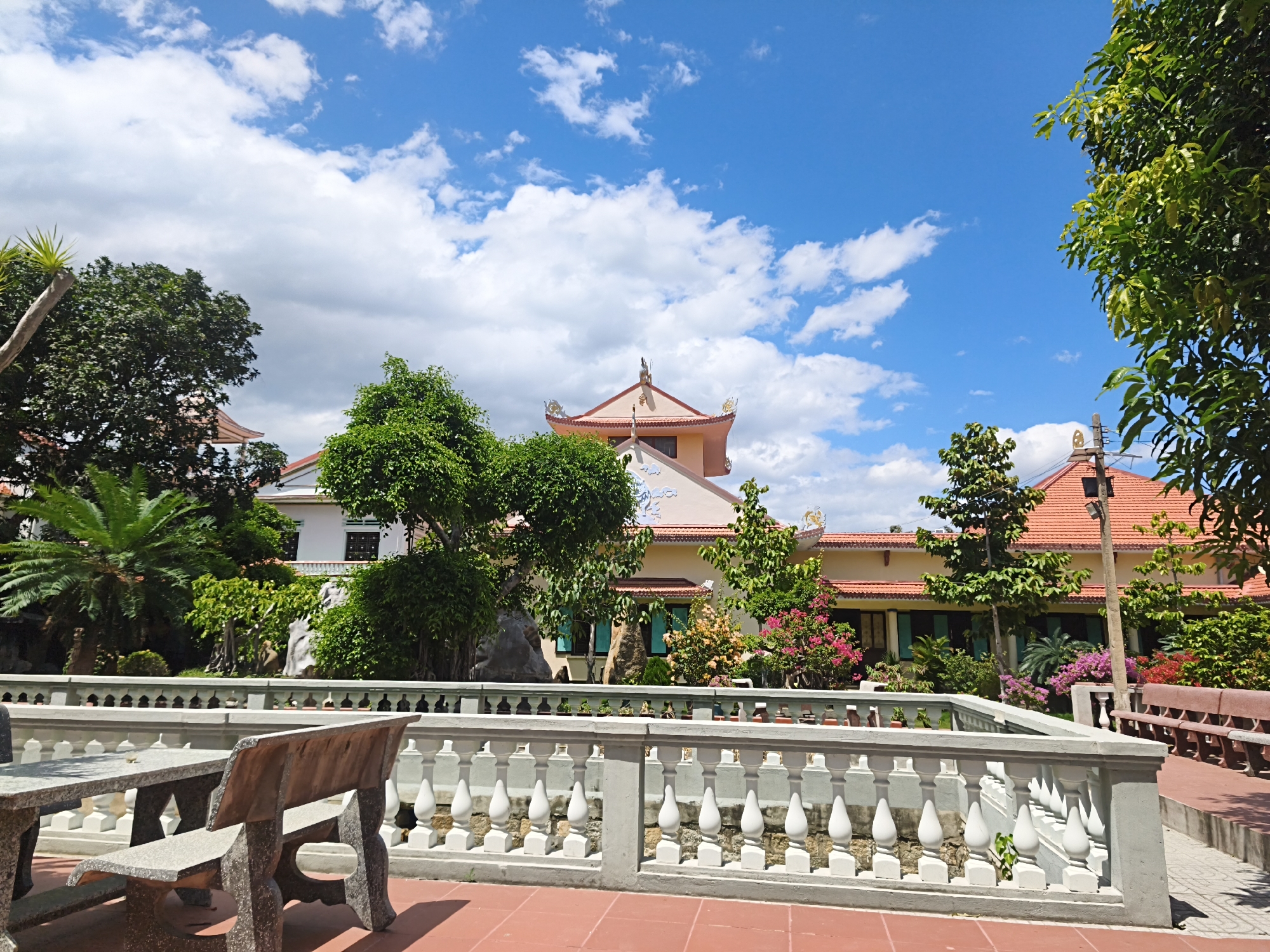 This is a matter-of-fact, forensic description of a courtyard area, likely at a religious site in Cam Ranh, Vietnam. In the foreground, a pink-tiled patio leads to a grey stone table and bench set, positioned in front of a white balustrade. The balustrade has numerous decorative white balusters and a grey concrete railing. Beyond the balustrade, lush green trees and flowering bushes create a vibrant garden. In the background, a light-colored building with an orange tiled roof is visible, featuring traditional East Asian architectural elements such as curved eaves and decorative finials. The sky above is a clear, bright blue with scattered white clouds, indicating fair weather. The scene appears to be serene and well-maintained, suggesting a peaceful atmosphere.