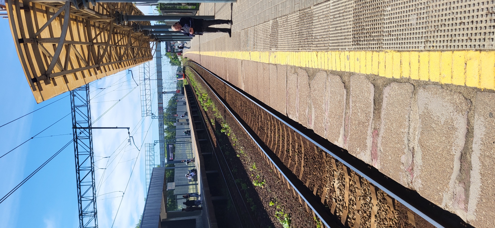 A man stands on a train platform, facing away from the camera, looking down at his phone. The platform is paved with light brown concrete blocks and has a yellow line marking the edge. Beyond the yellow line are train tracks with gravel ballast. A yellow awning, supported by metal poles, extends over the platform. In the distance, a red train is visible on the tracks, and other people can be seen on the platform. The sky is bright blue with a few wispy clouds, suggesting it is daytime and the weather is clear. The overall scene is a typical train station environment.