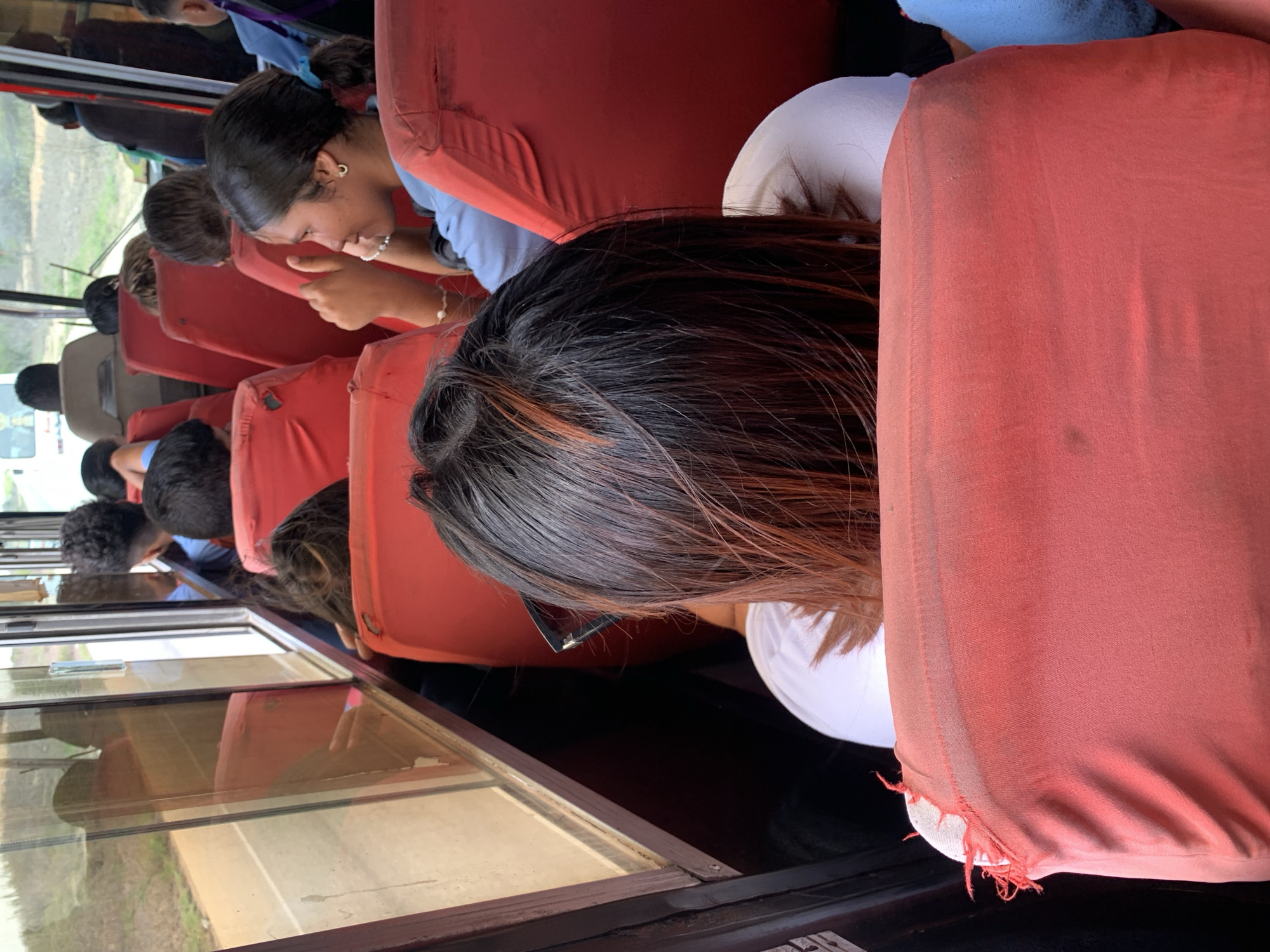 The image shows the interior of a bus traveling on a road near Santa María, Venezuela. The bus has red upholstered seats, and several passengers are visible. In the foreground, a person with long, dark hair with reddish highlights is seated, facing away from the camera, with their back to a red seat. To the right of this person, a young woman with dark hair tied in a ponytail is seated and looking to her right, giving a thumbs-up gesture. She is wearing a light blue shirt and pearl-like jewelry. Other passengers are visible further down the bus, with some looking out the window. The windows offer a view of the road and the surrounding rural landscape. The lighting suggests it is daytime.