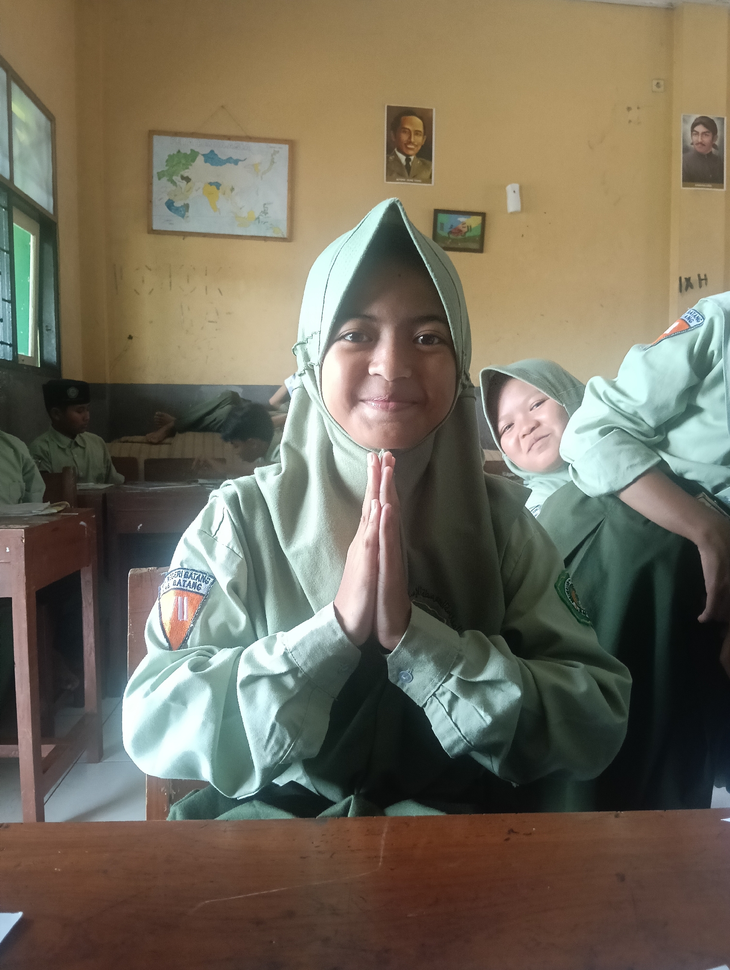 A group of young Indonesian students, likely in a classroom, are the subjects of this photograph. The central figure, wearing a light green hijab and school uniform, faces the camera with hands pressed together in a gesture of greeting or prayer. To her right, another student in a similar uniform smiles playfully. In the background, other students are visible, some seated at desks and one lying down. The classroom environment is evident with a world map and portraits on the wall, suggesting an educational setting. The room appears to be lit by natural light from a window on the left. The presence of the school uniform with insignia indicates this likely takes place within a school in Adinuso, Indonesia, which is known for its educational institutions.