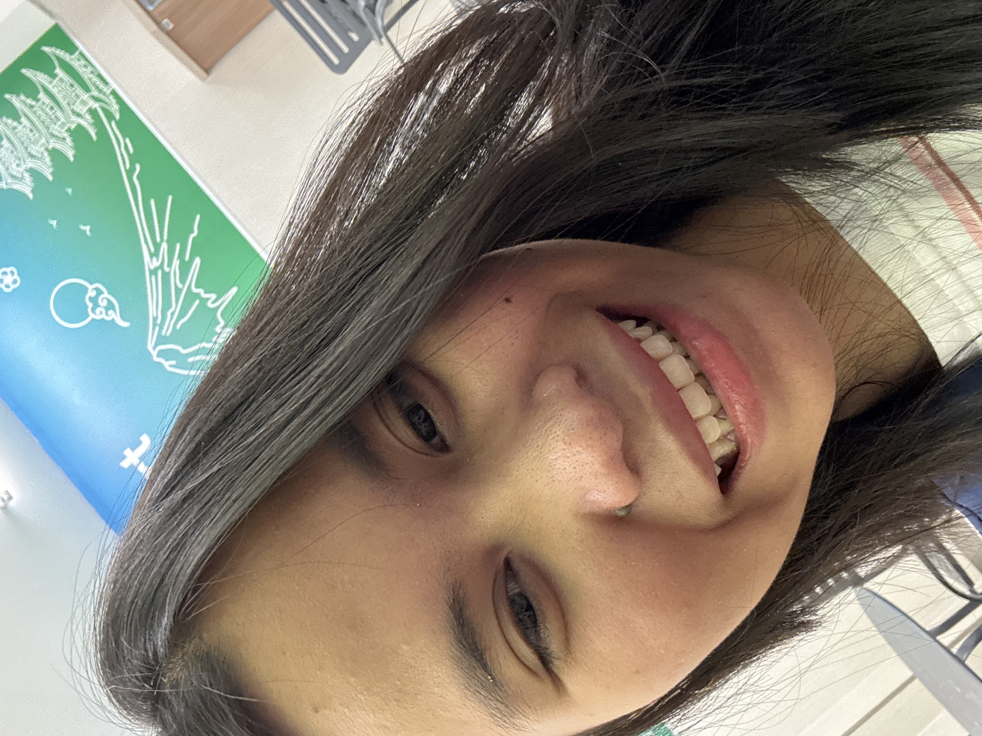 A close-up selfie of a young woman with long, dark hair smiling. She has dark eyes and her mouth is open, showing her teeth. Her skin appears to have some texture, with visible pores. She is wearing a white t-shirt with red and pink stripes. In the background, there is a mural on the wall depicting a mountain range with a pagoda and clouds, in shades of blue and green. Part of a white ceiling with lighting fixtures is visible at the top. To the right, a brown door and a portion of a gray chair can be seen. To the left, a gray table and chair are partially visible. The location is in East Jakarta, Indonesia.