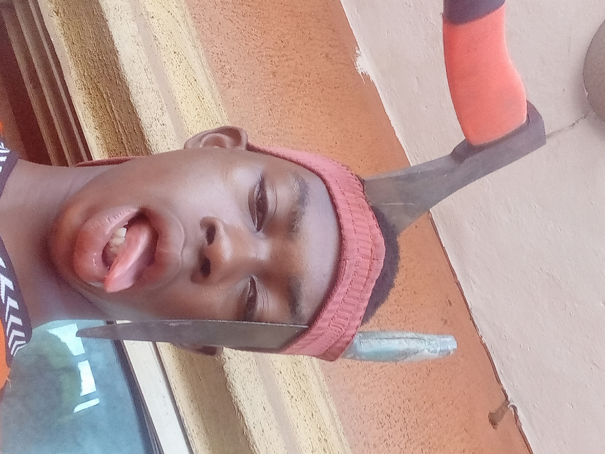 A young male with dark skin is seen in a close-up portrait. He has a red knitted headband. His mouth is open and his tongue is extended. There is a large metal object, possibly an ax, positioned above his head, and a metal knife is positioned under his chin. The background appears to be the walls of a building. The location is Nkpor, Nigeria.