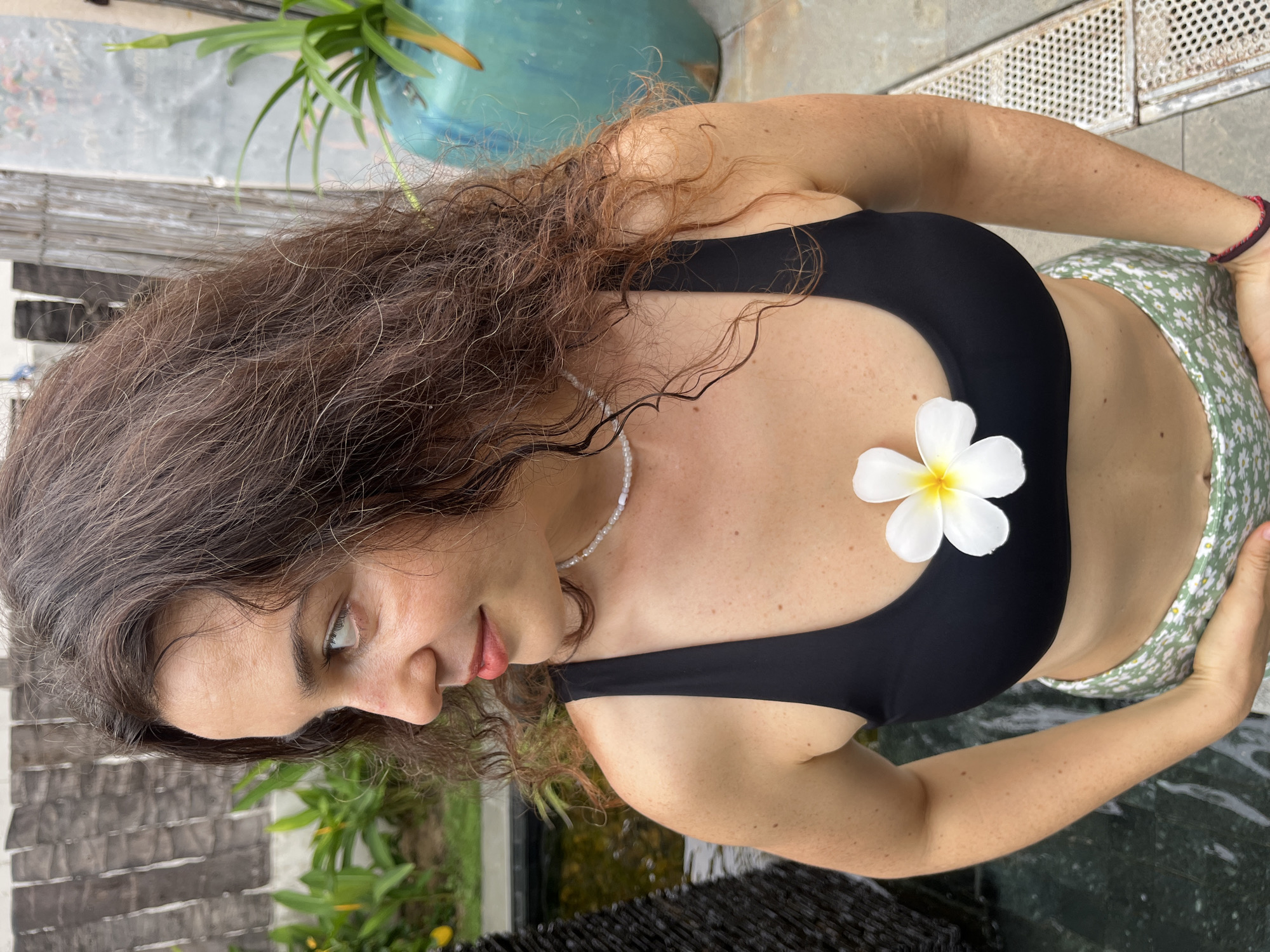 A woman is standing in what appears to be an outdoor space with a water feature and some greenery. She has long, wavy brown hair and is looking to her left. She is wearing a black bikini top with a white flower pinned to the left side of her chest. She is also wearing a green and white floral print skirt. A beaded necklace is visible around her neck. The background includes a dark, textured wall, lush green plants, and a large turquoise pot. There is also a glimpse of a water pool. The lighting suggests it is daytime. The location is identified as Phu Thuy Ward, Vietnam.
