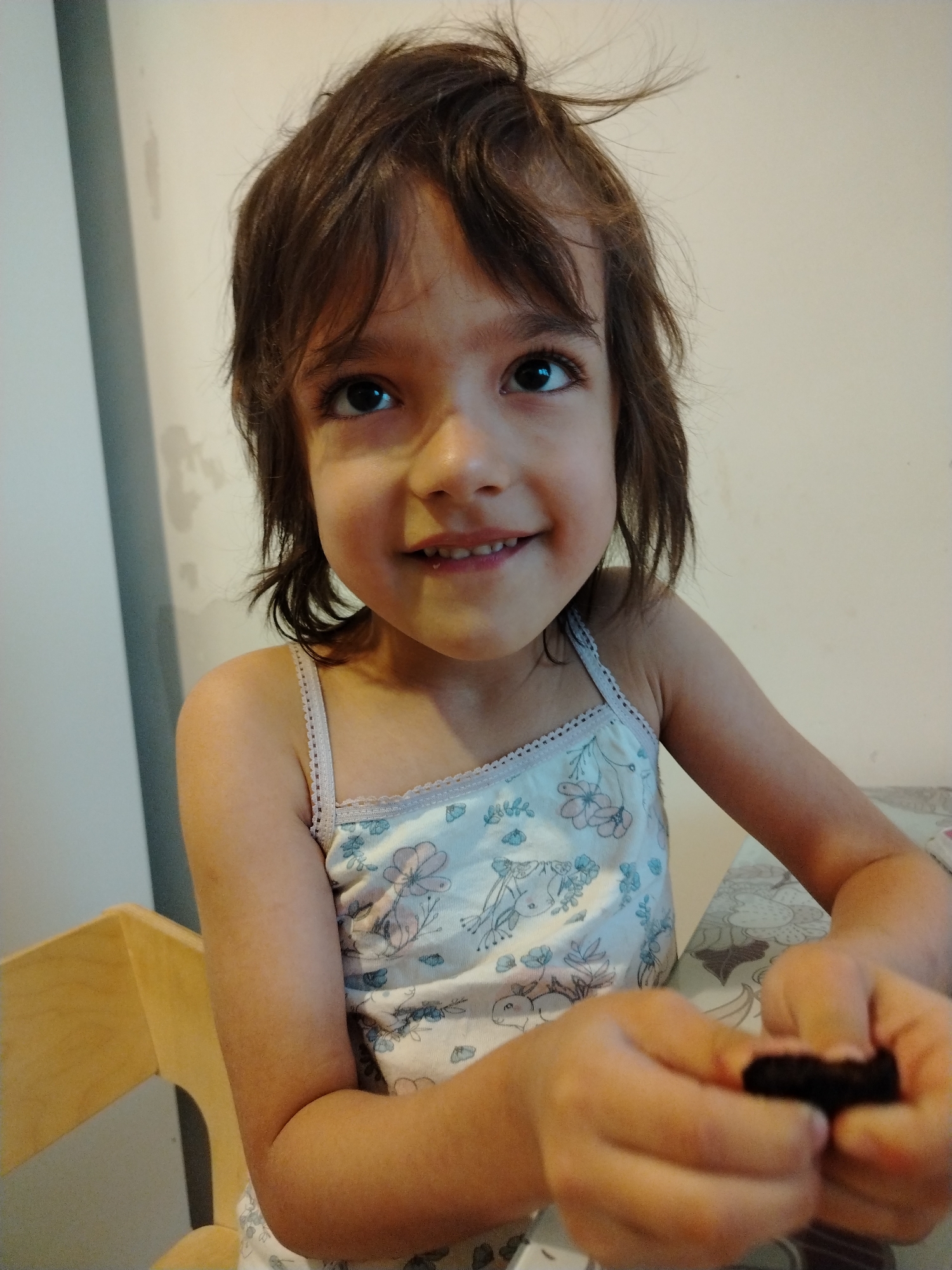 A young girl is seated at a table, holding a dark-colored food item (possibly a brownie or small cake) in her hands. She is wearing a light-colored tank top with a floral pattern. Her hair is brown and somewhat tousled. She is smiling and looking upwards. The background consists of a light-colored wall with some imperfections and a portion of a wooden chair is visible on the left. The tablecloth has a patterned design. The location in the city of Noviy, Russia is unknown.