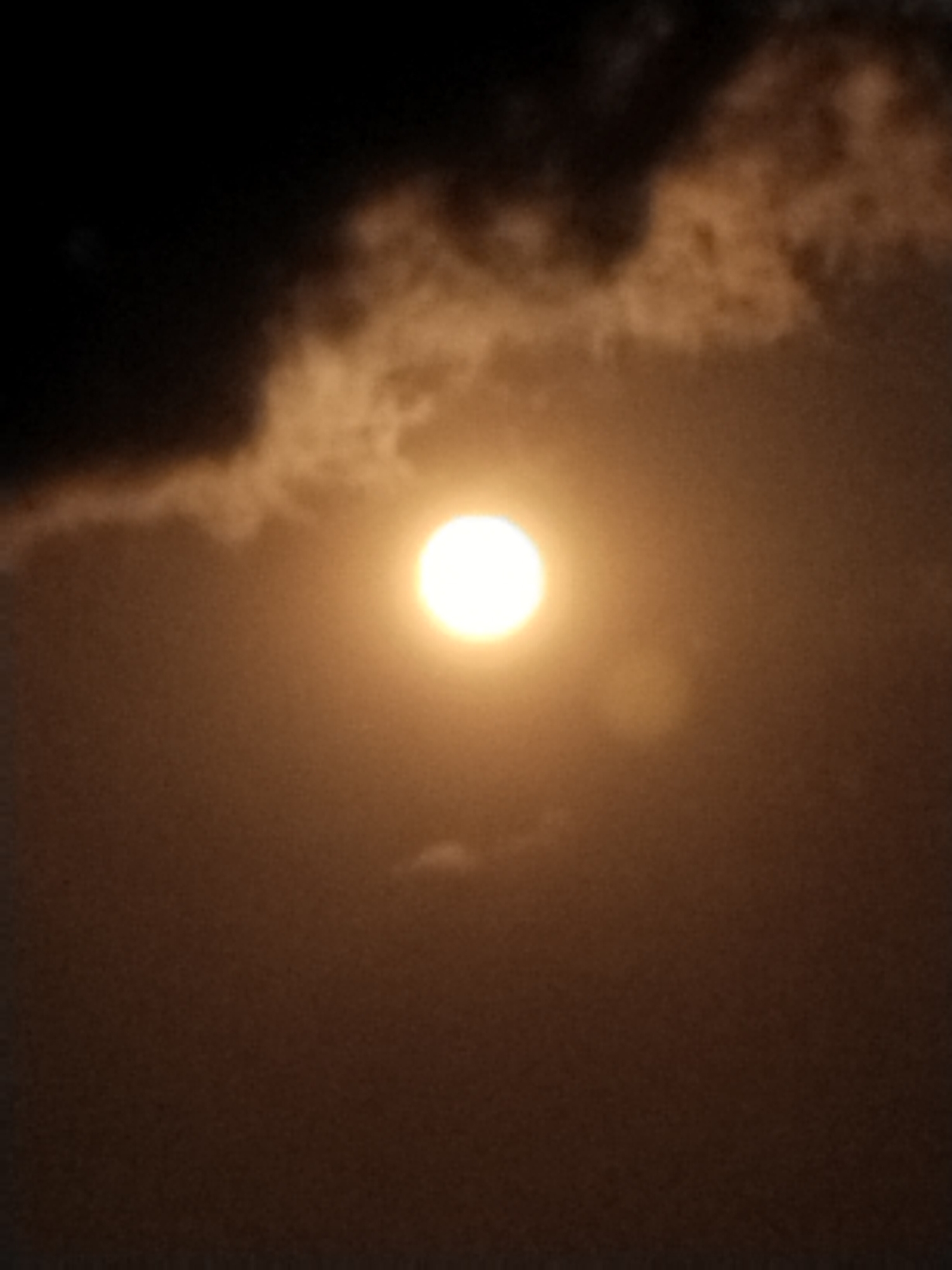 This is a photograph of the sun partially obscured by clouds, taken during the daytime in Gumeng, Indonesia. The sun appears as a bright, luminous orb in the center of the frame, surrounded by a hazy glow. Darker clouds are visible in the upper left quadrant, while lighter, more diffuse clouds are scattered across the sky. The overall color palette is dominated by warm oranges and browns, suggesting either a sunrise, sunset, or a heavily overcast sky. The image lacks any discernible human subjects, specific landmarks, or man-made objects that would indicate a precise location within Gumeng. The scene depicts a natural atmospheric phenomenon.
