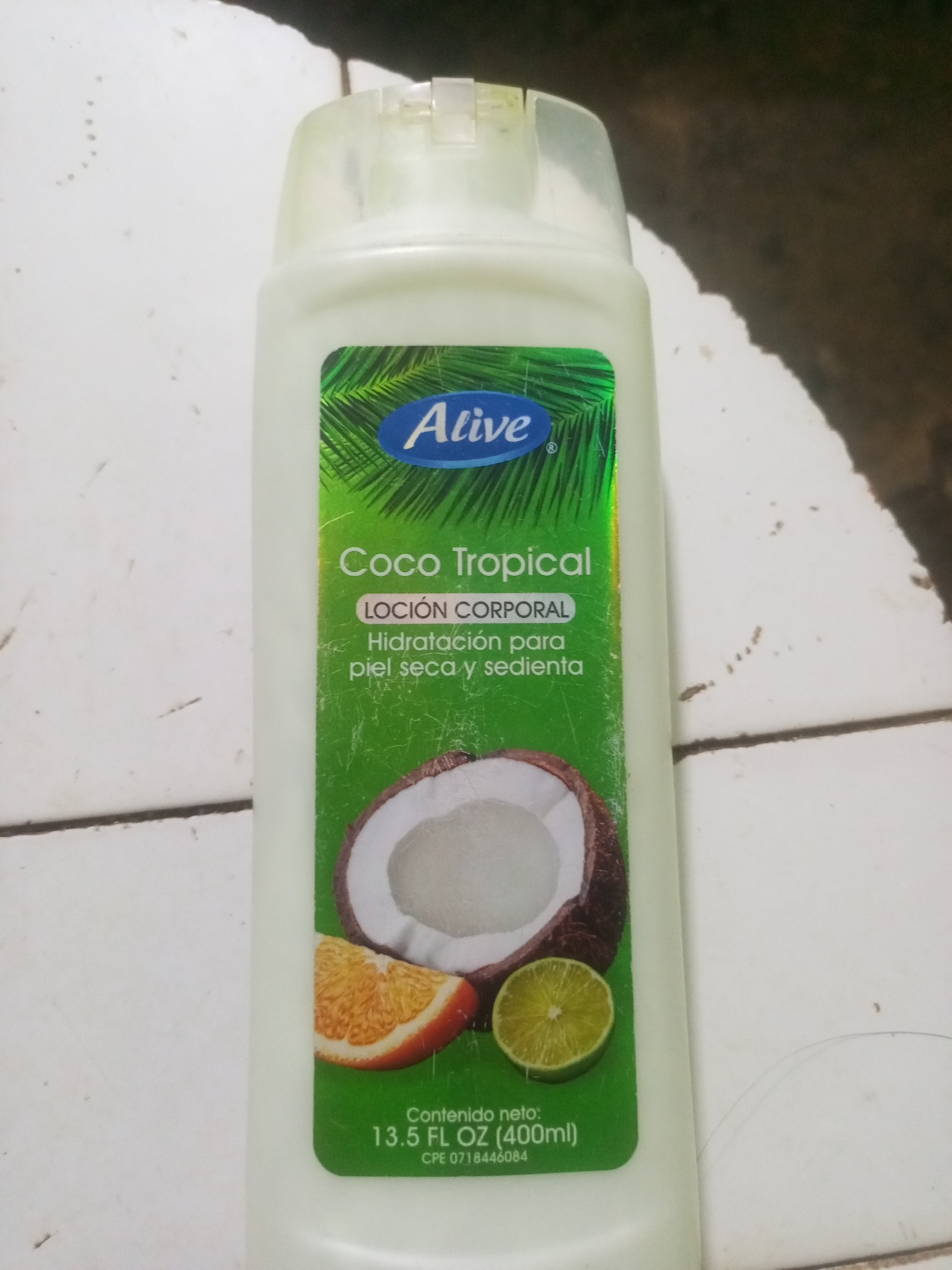 This is a close-up, high-angle, indoor, eye-level shot of a bottle of lotion. The bottle is white and opaque, with a clear plastic flip-top cap. The label on the bottle is green with a holographic sheen. The label features the brand name "Alive" in a blue oval, followed by "Coco Tropical" in white text. Below that, in smaller text, it reads "LOCIÓN CORPORAL Hidratación para piel seca y sedienta." The label also includes an illustration of a halved coconut, a slice of orange, and a slice of lime. At the bottom of the label, it states "Contenido neto: 13.5 FL OZ (400ml)" and a product code "CPE 0718446084". The bottle is placed on a white surface with a subtle texture and faint grey lines, possibly a tiled floor or countertop. The background is out of focus and appears to be a dark, textured surface. There are no people or animals visible in the image. The lighting is artificial, creating some glare on the plastic cap. The image does not provide any information to suggest it is located in La Tagua, Venezuela.