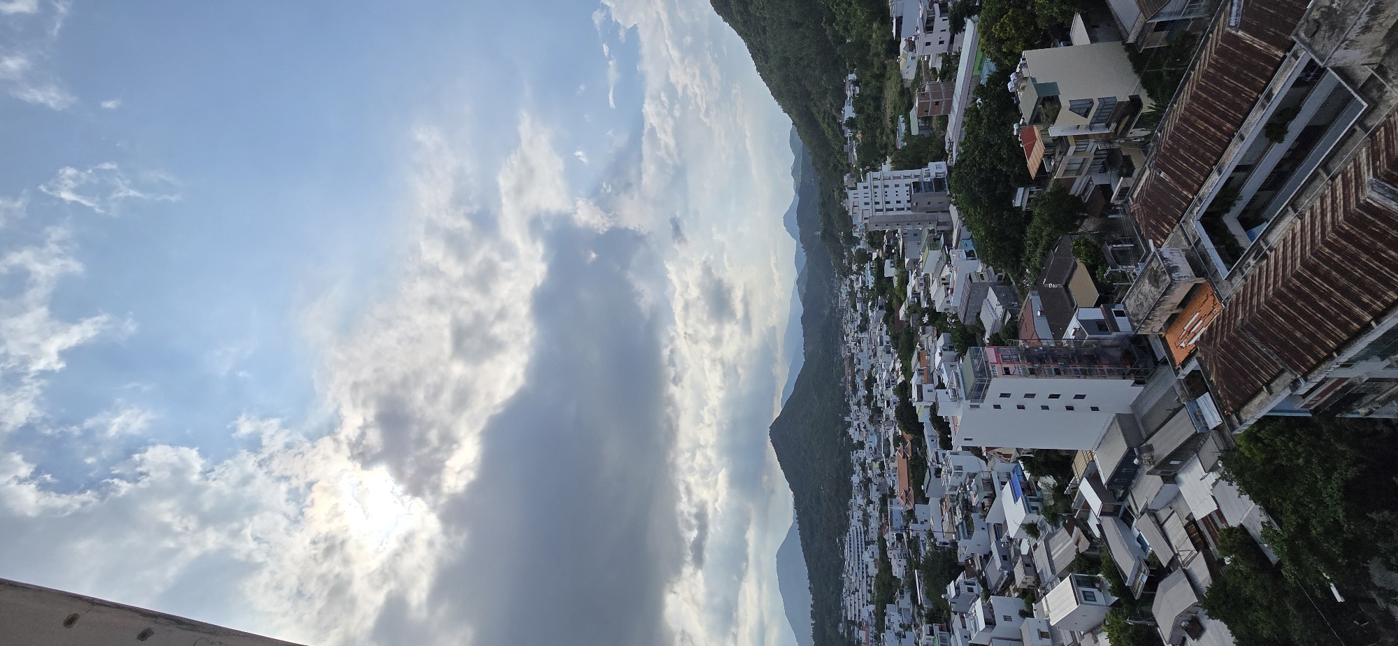 This is a high-angle, wide shot of a city overlooking a bay in Nha Trang Ward, Vietnam. The sky is partly cloudy, with the sun peeking through the clouds on the left side of the frame, creating a bright halo effect. The city below is densely populated with buildings of various sizes and colors, ranging from small, low-lying houses to taller, multi-story structures. Many of the roofs are corrugated metal, some showing signs of rust. Green trees and vegetation are interspersed throughout the urban landscape, with lush, tree-covered hills forming a backdrop to the city. Several mountain ranges are visible in the distance, fading into a hazy blue. The overall impression is of a vibrant, bustling city nestled within a tropical environment. There are no people or obvious activities visible in the image.