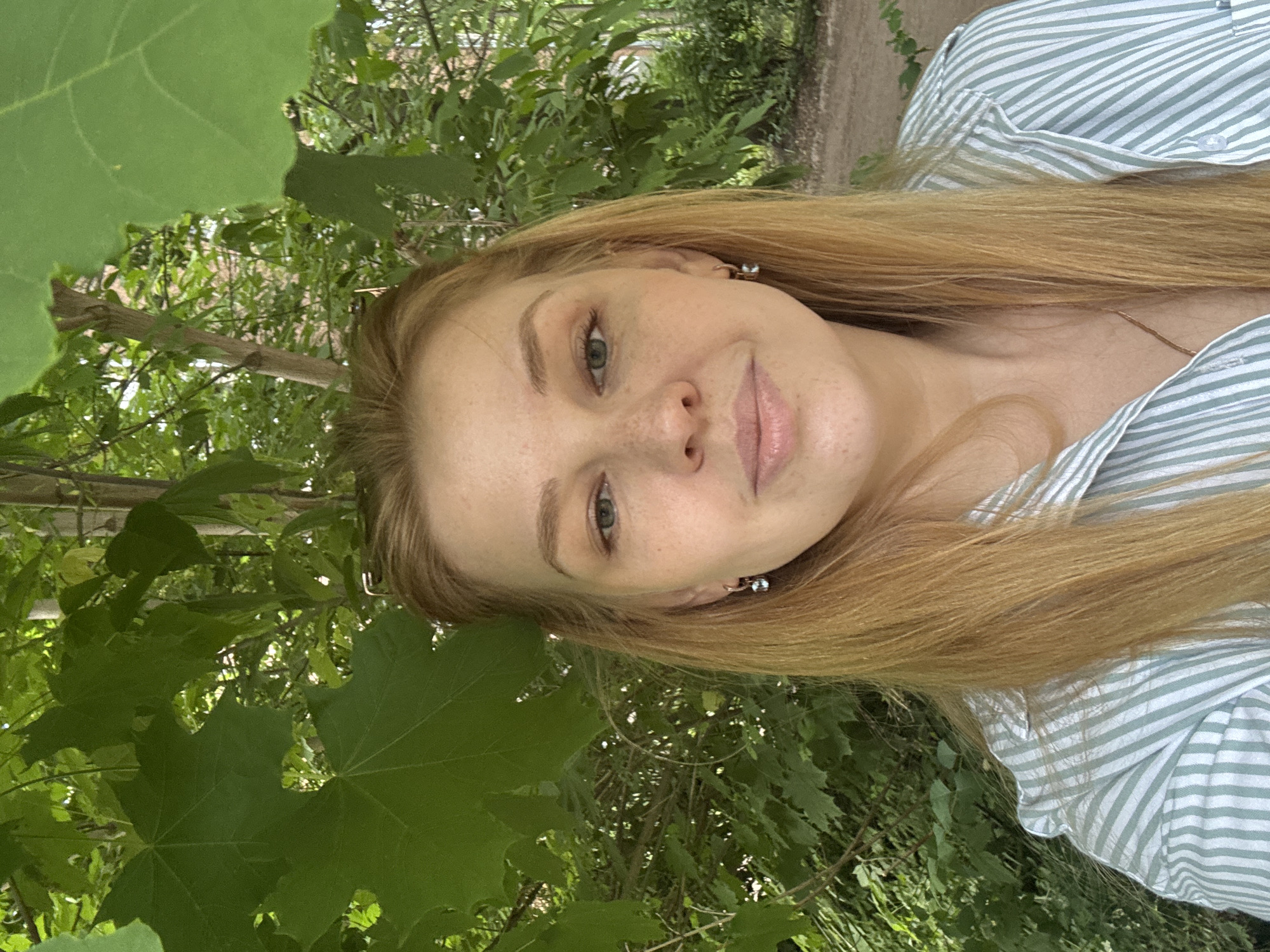 This is an outdoor portrait of a young woman in a natural setting, likely a park or garden in Rusanovo, Russia. The woman, with long, straight, light brown or blonde hair, is positioned in the center, looking directly at the camera. She displays a subtle, closed-mouth smile, conveying a calm and content expression. Her eyes appear to be a light color, possibly green or hazel, and her face is dotted with numerous small freckles across her nose and cheeks.
She is wearing a light-colored collared shirt, featuring thin, light green vertical stripes. Small, silver-toned earrings are visible, and a delicate gold-colored chain necklace rests around her neck.
The background is dominated by a dense array of lush green foliage, consisting of various leaves and branches from trees, indicating a vibrant, verdant environment. The lighting suggests a bright, sunny daytime, with natural light illuminating the scene and dappling through the leaves. There is no visible text in the image.