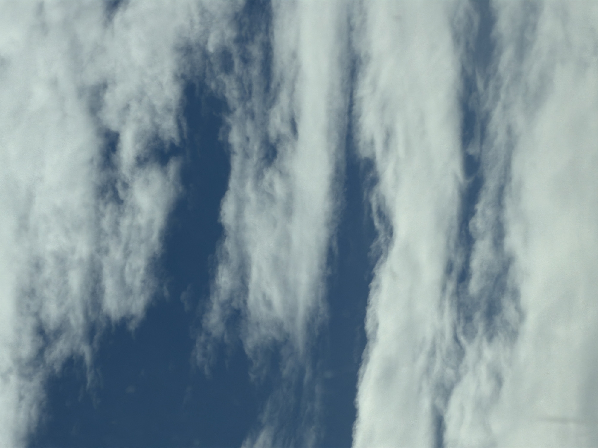 This media file presents a close-up, abstract view of the sky over North Kuta, Indonesia. The primary subjects are wispy white clouds set against a vibrant, deep blue sky. The clouds are predominantly elongated and appear to stretch vertically across the frame, creating dynamic, streaky patterns.
Some cloud formations are denser and more opaque, forming broad bands of white, while others are thinner and more translucent, allowing the underlying blue sky to peek through. Distinct vertical channels of clear blue sky are visible between the cloud masses, highlighting the contrast between the two elements. The texture of the clouds varies from soft and ethereal to slightly more defined, with subtle swirling patterns visible in places.
The lighting suggests it is daytime, with soft and even illumination across the sky and clouds, indicating no direct harsh sunlight or shadows. No specific activity or event is taking place, as the image focuses solely on the natural atmospheric conditions. There are no people, man-made objects, or visible text within the frame. The overall impression is one of natural beauty, vastness, and the ever-changing patterns of the sky.