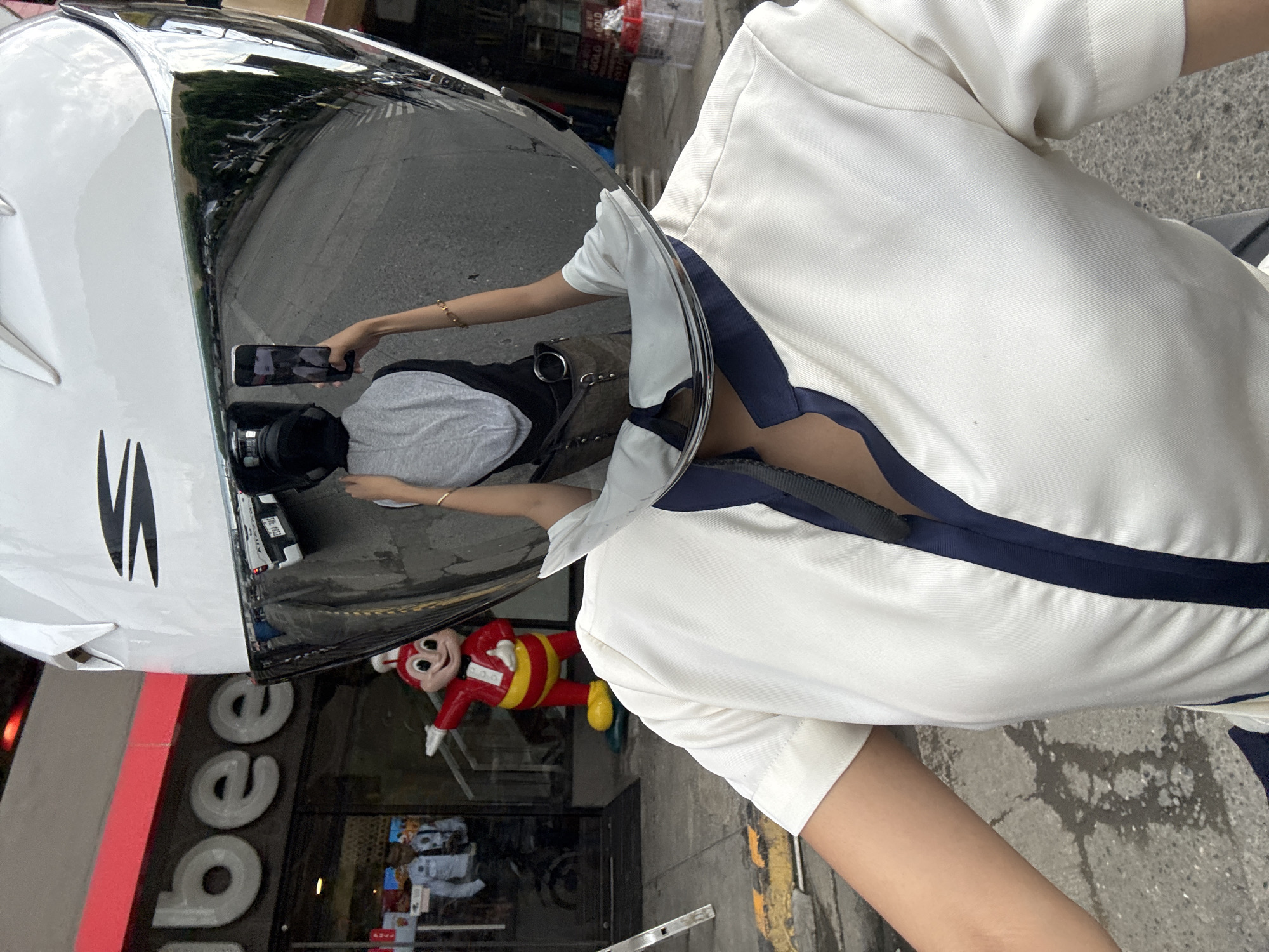 The image shows a person wearing a white helmet with a reflective visor, obscuring their face. The visor reflects a street scene, including a Jollibee restaurant with its iconic mascot. In the reflection, a person is visible holding up a smartphone to take a selfie. The foreground shows the person's torso and arm, wearing a white shirt with dark blue trim. The background outside the restaurant appears to be a typical street in Davao City, Philippines, with vehicles and pedestrians.