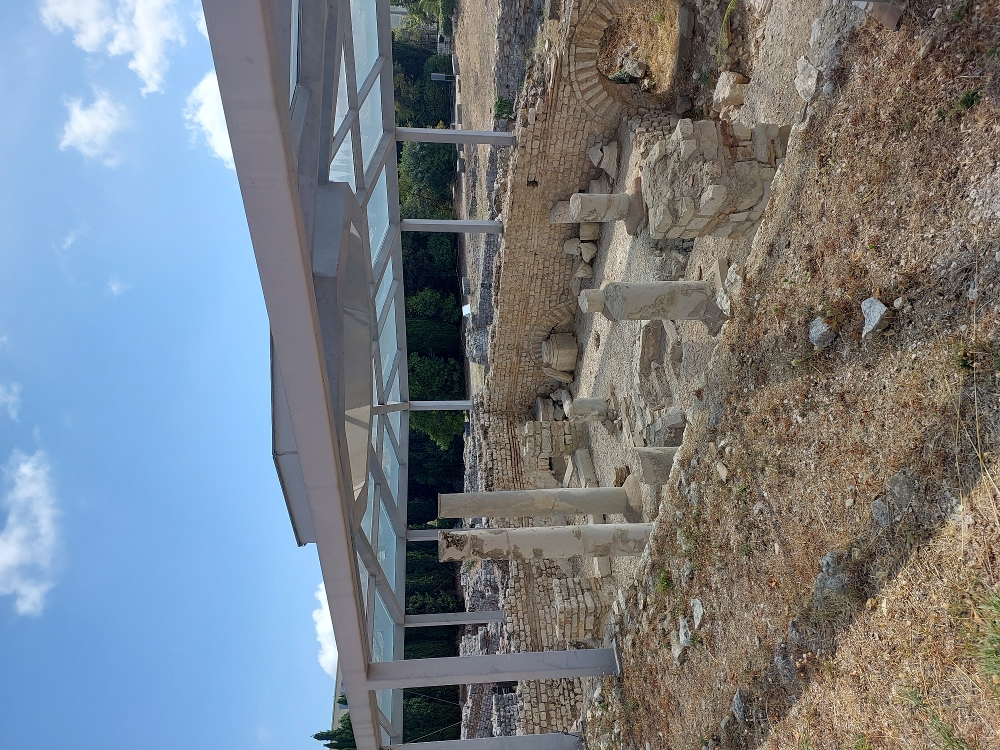 The image depicts an archaeological site in Nice, France, featuring the ruins of ancient structures. A modern protective canopy with a glass roof and metal supports is erected over a portion of the ruins, shielding them from the elements. The ruins consist of stone walls, partially collapsed columns, and arched structures, indicative of historical buildings. The ground is covered with gravel and dry vegetation. In the background, a line of trees forms a natural border, and beyond them, a modern building is visible. The sky is blue with scattered white clouds. No people are present in the image.