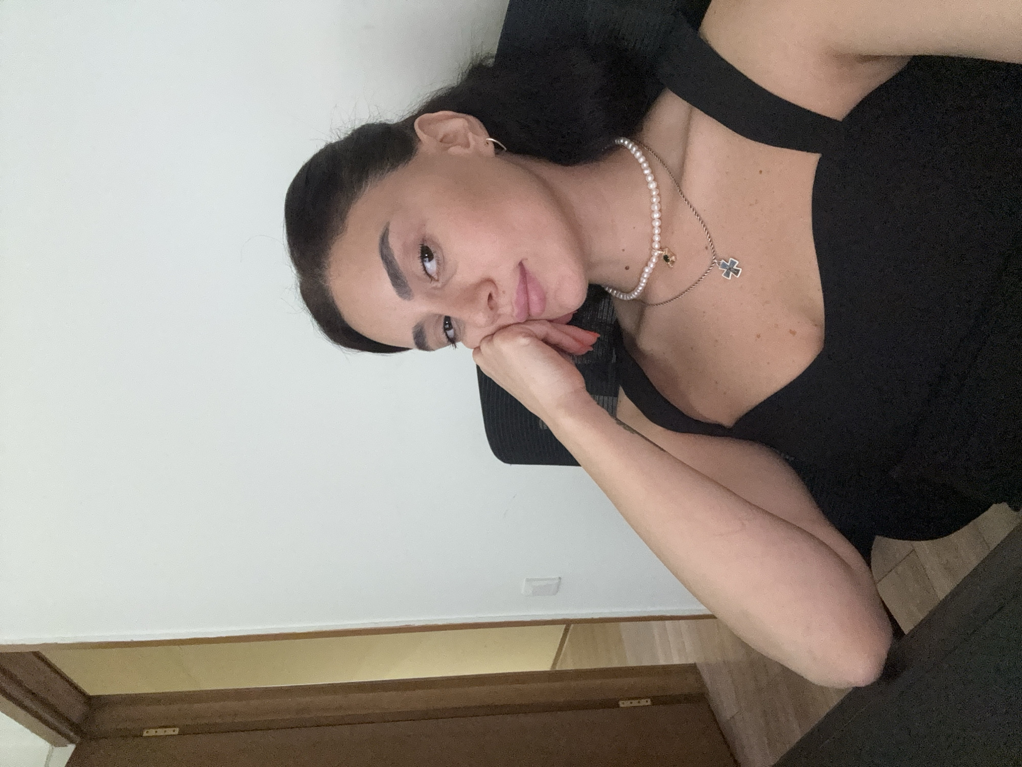 A young woman with dark hair is sitting in an office chair, leaning her head on her hand. She is wearing a black top and two necklaces, one of which is a pearl choker. The background shows a white wall and a doorway leading to another room. The scene appears to be indoors, possibly in an office or home environment. There are no specific details in the image that indicate the location in Tbilisi, Georgia.