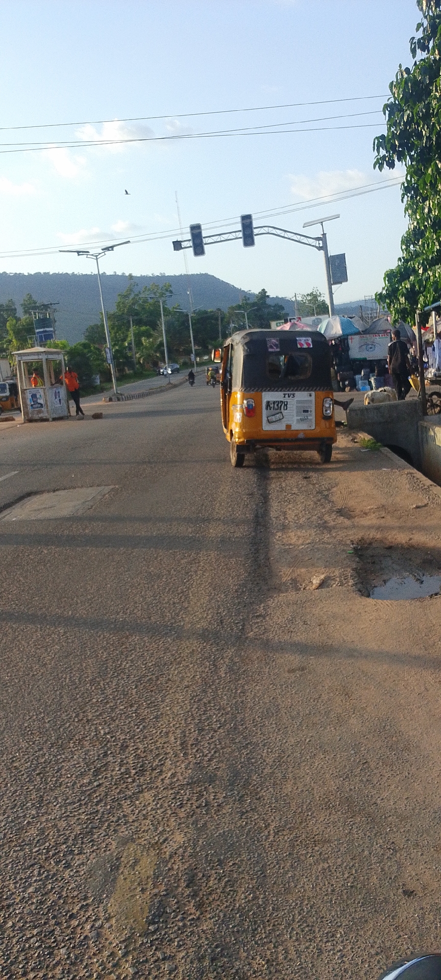 The image depicts a street scene in Lokoja, Nigeria, under a bright sky with a visible bird. A yellow and black "keke napep" (auto-rickshaw) is the central subject, facing away from the viewer and displaying a license plate with "K-1378."
The street surface appears rough and textured, with possible patches or repairs. To the right, a raised curb with a puddle near the edge borders a scene of market stalls or roadside vendors, with people standing near them. In the distance on the left, there's a small kiosk with an individual in an orange shirt standing next to it. Street lights and traffic signals are visible overhead, suggesting a main road or intersection. A hill can be seen in the background. There are cars and motorcyclists further down the road.