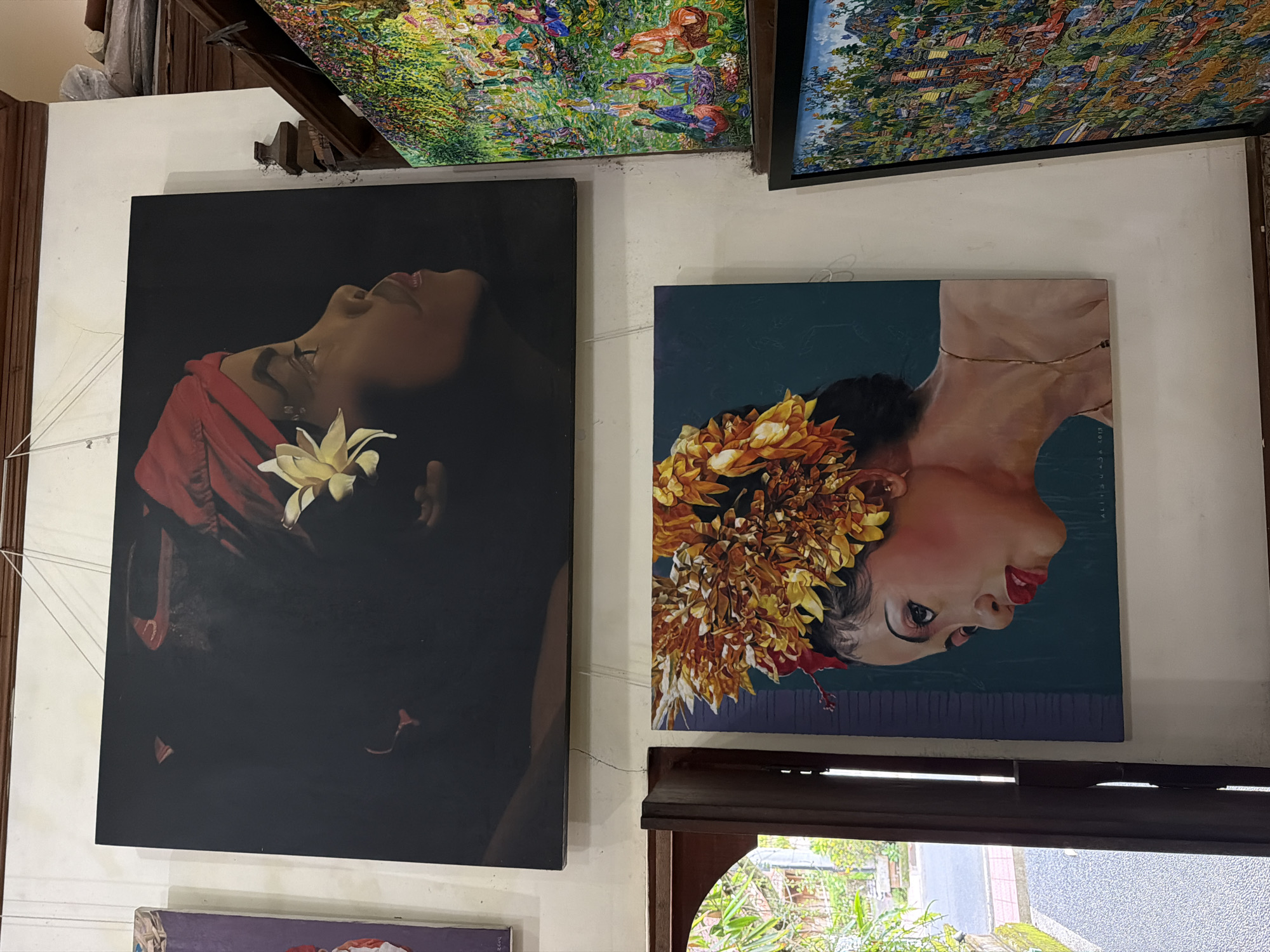 The image shows a collection of paintings on a wall in what appears to be a gallery or shop in Ubud District, Indonesia. Two large, framed paintings of women are prominently displayed. The left painting features a woman with dark hair, a red garment, and a white flower. The background is dark. The right painting depicts a woman with a floral headdress against a greenish-blue background. The paintings are hung slightly off-center. Other framed paintings are visible above, partially out of focus. The wall is off-white with some minor cracks and imperfections. A window is visible at the bottom, revealing a view of lush greenery outside. A wooden window frame and blinds are in the foreground. The composition suggests an interior setting with artistic displays.
