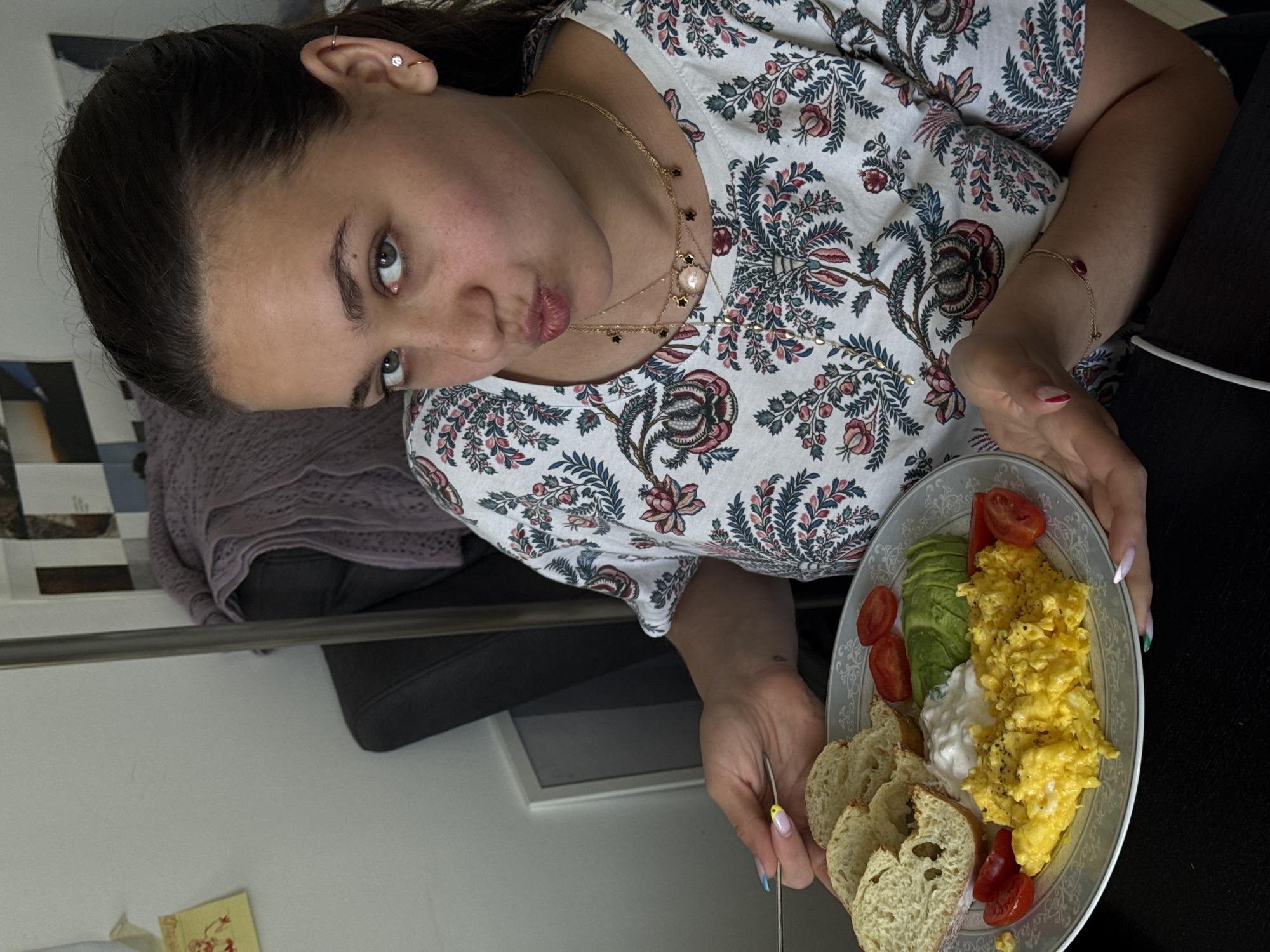 A young woman with dark hair and light eyes is pictured indoors, looking directly at the camera with a slight pout. Her dark hair is pulled back, revealing multiple piercings on her right ear, including a small hoop and a stud with a star or flower design. She is wearing a white short-sleeved top with a detailed floral and paisley pattern in red, blue, green, and black. Around her neck, she has layered gold necklaces, one with a central round pendant and another with smaller charms. A delicate gold bracelet with a red bead adorns her right wrist.
She holds an oval plate with both hands, showcasing a well-presented meal. The plate contains a substantial serving of scrambled eggs, several slices of ripe avocado, halved cherry tomatoes, a dollop of white creamy substance (likely yogurt or cream cheese), and several pieces of rustic-looking bread. Her nails are manicured, with red tips on her right hand and distinctive yellow and blue tips on her left hand, which also holds a fork.
The background suggests an indoor setting, possibly a home. A dark-colored sofa or chair is visible, draped with a folded purple knitted throw. The lighting appears natural and bright, indicating it is likely daytime. The overall impression is that of a person enjoying a prepared meal, possibly breakfast or brunch, and posing for the photograph. No explicit text is visible in the image.