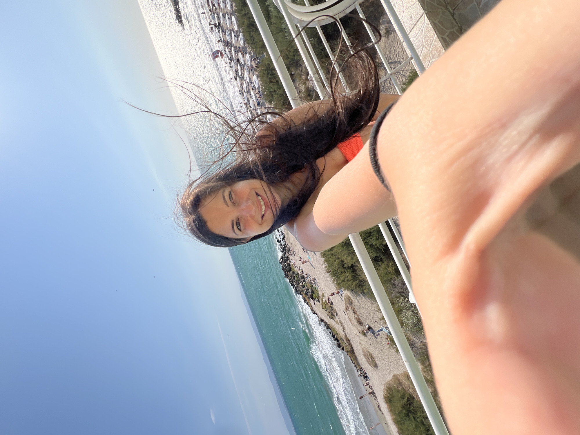 A woman is taking a selfie on a balcony overlooking a beach in Pomorie, Bulgaria. The woman has long, dark hair that is blowing in the wind. She is smiling and wearing an orange bikini top. The beach is crowded with people sunbathing and swimming. The sea is a vibrant turquoise color, and the sky is a clear blue. There are umbrellas and lounge chairs on the beach. The balcony railing is white. The image is taken from a high angle, looking down at the beach.