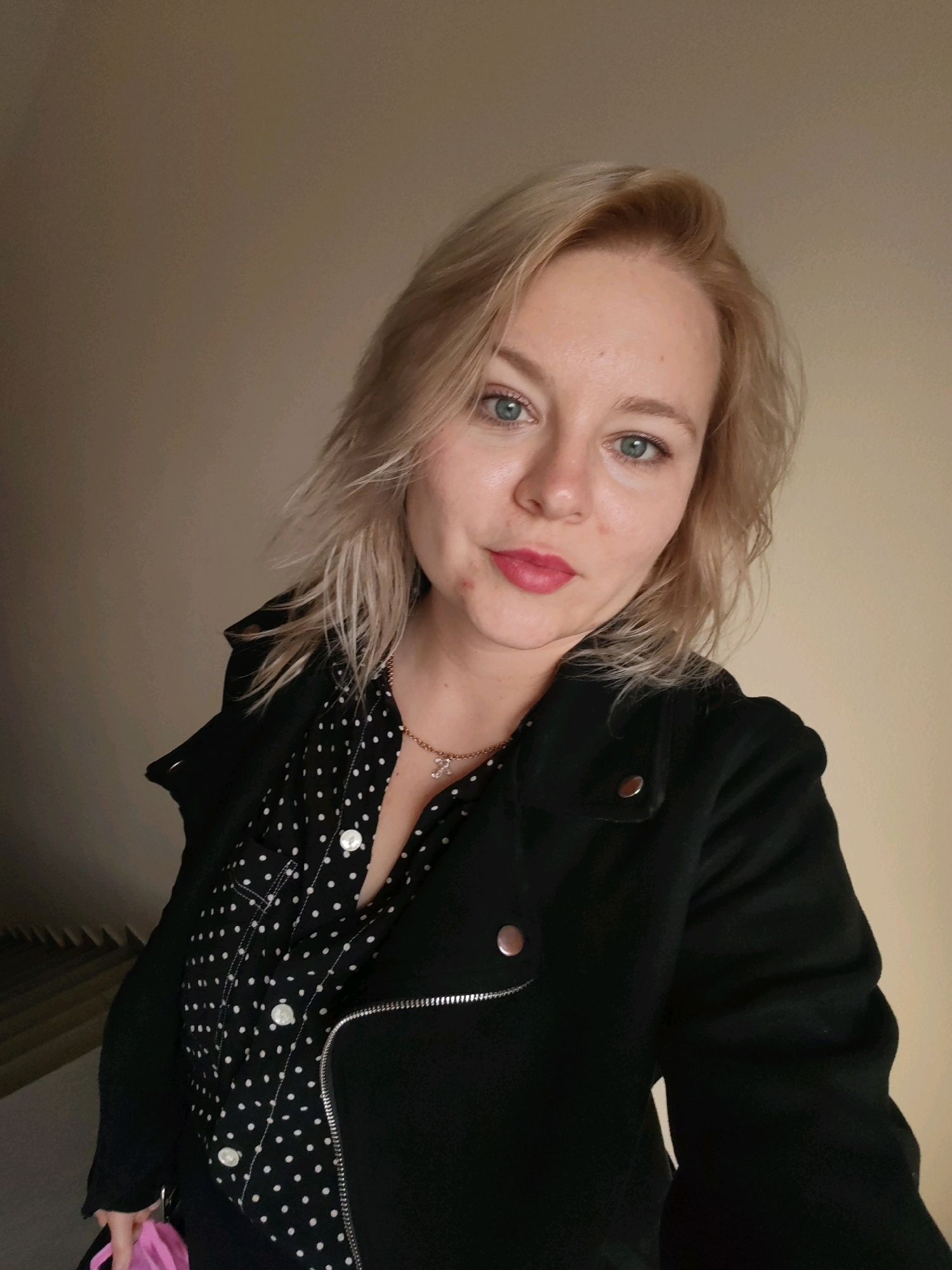 A woman with blonde hair and blue eyes, wearing a black jacket over a black and white polka dot shirt and a necklace, stands in front of a beige wall. Her head is tilted slightly, and she is looking directly at the camera. A pink bag is visible in her left hand. The background suggests an indoor setting, possibly a hallway or corridor, with a staircase partially visible on the left. There is no specific information in the image to place this scene within the city of Izhevsk, Russia.