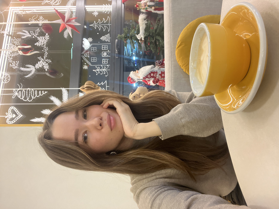 The image displays a young adult female seated at a light-colored table within an indoor establishment, likely a cafe, adorned with Christmas decorations. The specific location within the city of Krasnoyarsk, Russia, cannot be determined from the visual evidence.
The subject has long, straight brown hair and is looking directly at the camera with a neutral expression, her lips slightly parted. She is wearing a light beige or tan turtleneck sweater with ribbed cuffs. Her left hand is positioned under her chin, and a small hoop earring is visible on her left ear.
On the table in the foreground, a bright yellow ceramic coffee cup containing a light brown beverage with foam is placed on a matching yellow saucer. Behind the subject, a large window is decorated with white chalk-like drawings depicting various Christmas elements, including hearts, hanging ornaments, mistletoe, houses, and pine trees. Several physical decorations are also present: hanging wooden or paper star ornaments in red and white, and a brown honeycomb-style ornament. On a ledge behind the window, visible figurines include a Santa Claus, a small artificial Christmas tree with a white squirrel ornament, and a Nutcracker. The view through the window shows blurred artificial lights, suggesting an urban environment at night. A light-colored sofa with a yellow cushion is partially visible behind the table.