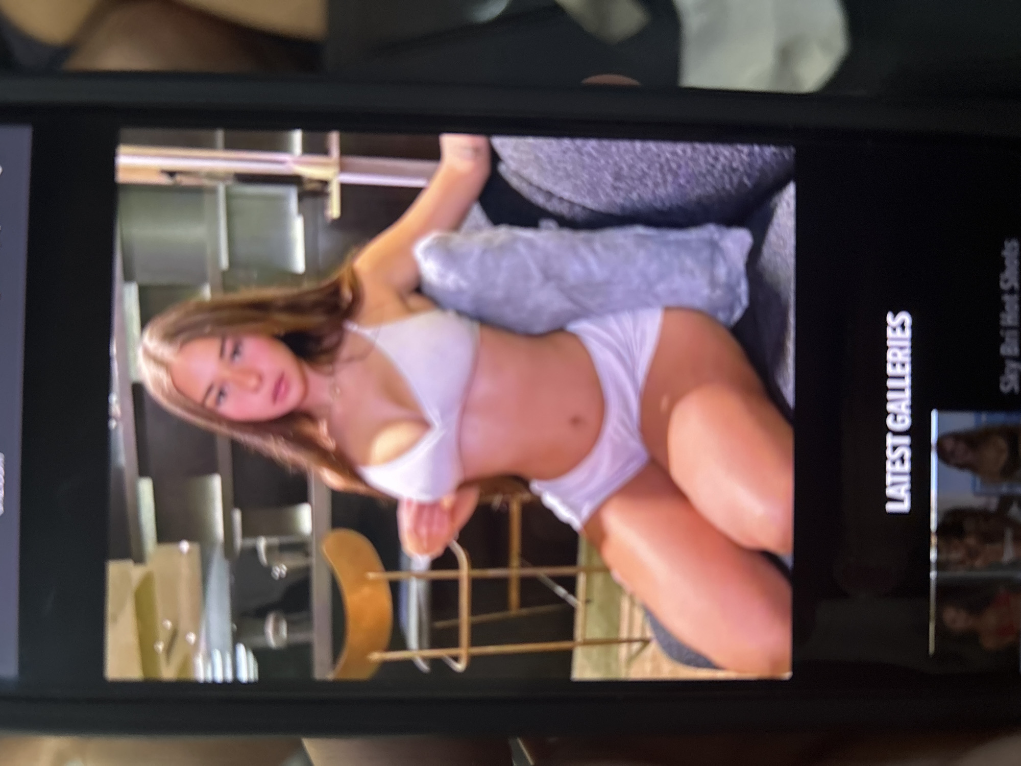 A photograph captures a smartphone screen displaying a digital image of a young woman. The woman, with long brown hair, fair skin, and a neutral expression, looks directly forward. She is wearing a white bralette and white shorts, seated with her legs extended. Her left hand is raised, appearing to adjust the strap of her bralette, while her right arm rests on a textured grey cushion or pillow. The background within the digital image shows an indoor setting with a light brown wooden bar stool to the left and modern shelves or architectural details behind her. Below the main image on the phone screen, white text reads "LATEST GALLERIES," and further below, partially visible text states "Sky Bri Hot Shots," next to three small thumbnail images. The phone itself is black, and portions of a hand holding the device are visible on the left and right edges of the overall photograph. There is no visual information within the image to indicate a location in Blagoevgrad, Bulgaria.