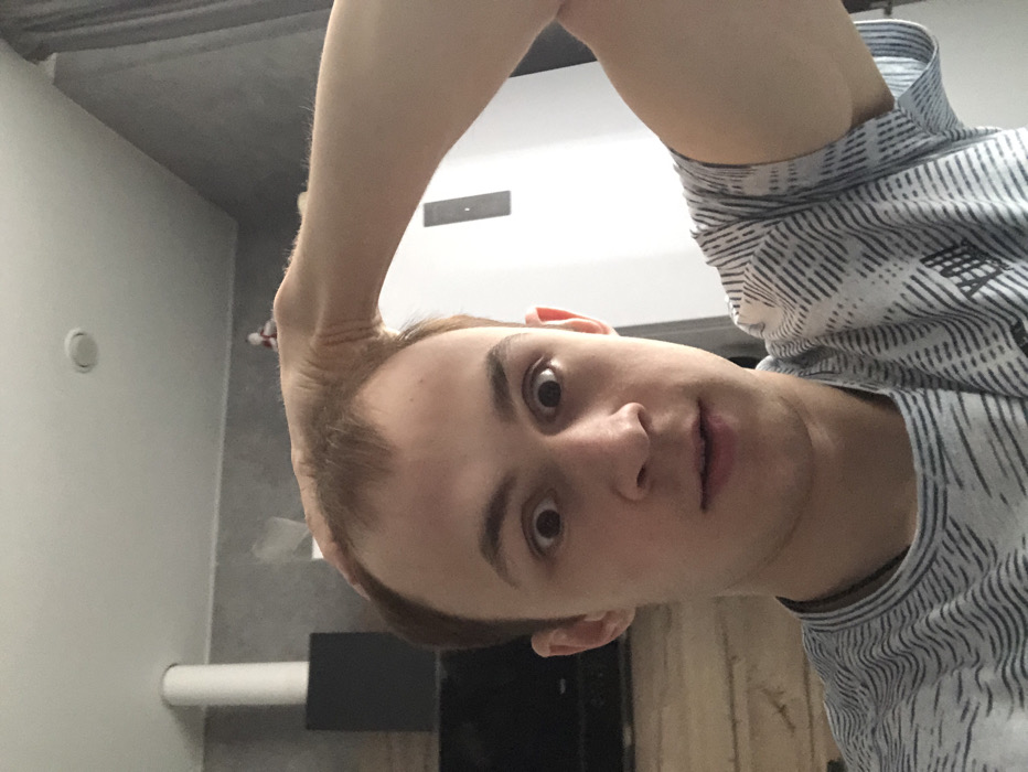 The image depicts a young adult male indoors, situated in what appears to be a residential environment within the city of Krasnodar, Russia. The male, with fair skin and light-colored, receding hair, occupies the central foreground, looking directly at the camera with a wide-eyed expression and slightly parted lips. His left hand (viewer's right) is positioned on his forehead, near his hairline. He is attired in a light-toned t-shirt featuring a dark, repeating wavy line pattern. The background on the left includes a dark, rectangular object resting on a wooden surface, behind which a white cylindrical element rises towards the ceiling. The wall behind him is a textured light grey. To the right, a white panel or surface with a dark vertical handle is visible, alongside grey curtains. A circular recessed light fixture is present in the white ceiling.
