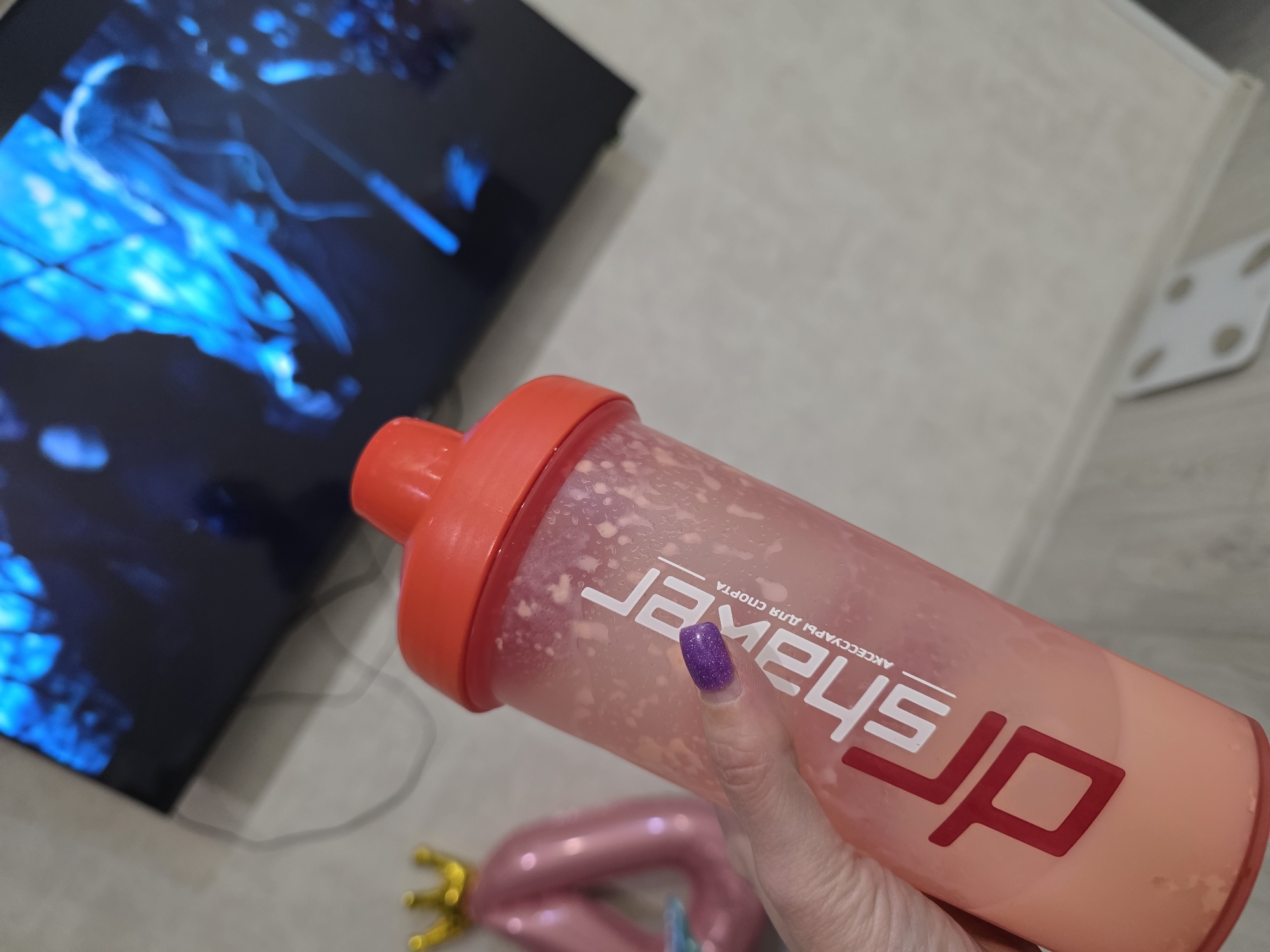 The image presents a close-up indoor scene, dominated by a hand holding a shaker bottle. The bottle is translucent pinkish-orange with a vibrant red lid, containing a frothy, light-colored liquid with small white particles, consistent with a protein shake or similar beverage. A hand, with fingernails painted in iridescent purple glitter polish, grips the bottle.
Prominently featured on the bottle in white text is the brand name "drSHAKER," oriented vertically. Below this, in smaller white letters, is the Russian phrase "АКСЕССУАРЫ ДЛЯ СПОРТА," which translates to "ACCESSORIES FOR SPORT."
In the background, to the left, a flat-screen television displays an abstract pattern of bright electric blue light against a dark screen. The setting appears to be a home, with a light-colored tiled or laminate floor. Partially visible at the bottom of the frame are a small, golden, crown-shaped object and a pink inflatable object, possibly shaped like a "0" or "O." To the upper right, a white body composition scale or similar floor-standing device with four circular elements is faintly discernible on the floor.
The scene captures a casual moment, likely related to personal wellness or relaxation at home. The provided location context is Chaikovsky, Russia.