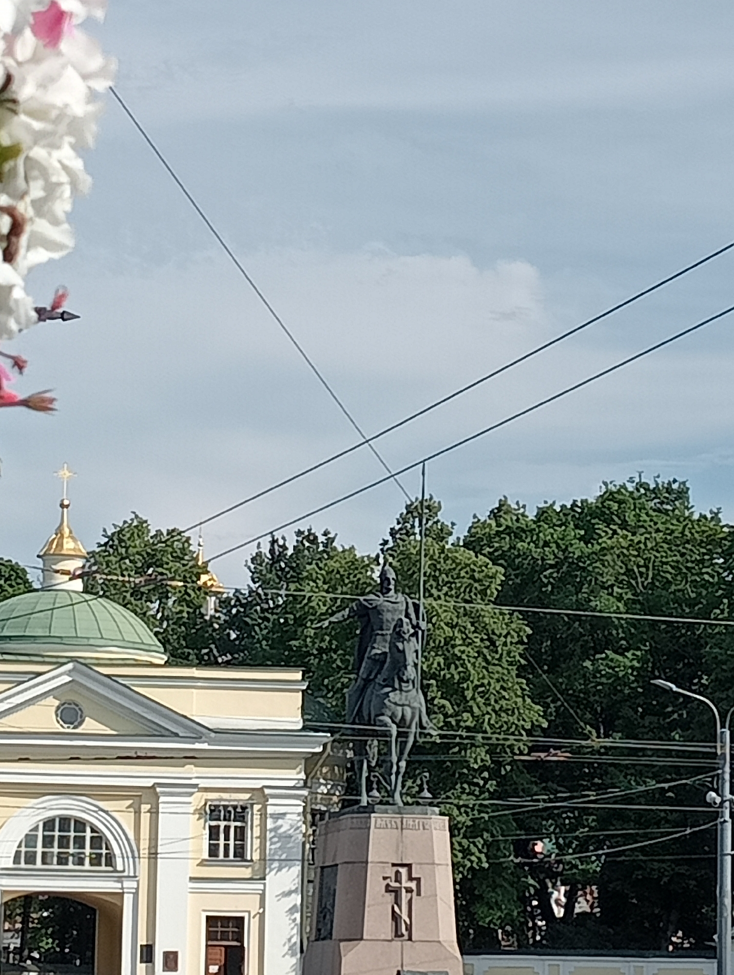 A statue of a mounted figure, likely a historical leader, stands prominently in an urban setting. The statue is made of dark metal and depicts the figure on horseback, wearing armor and holding a sword aloft. It is mounted on a tall, stone pedestal adorned with a large cross.
In the background, a light yellow building with white architectural details and a green dome is visible. A golden cupola with a cross tops a smaller structure near the building. Lush green trees form a dense backdrop. Several power lines crisscross the sky above the scene.
In the foreground, the edge of a blooming plant with white and pink flowers is visible, adding a touch of nature to the urban landscape. The sky is partly cloudy, suggesting it could be a partly sunny day. The overall impression is one of historical significance and civic pride, set against a backdrop of architecture and nature. The time of day appears to be daytime, given the lighting conditions. There is no discernible text in the image.