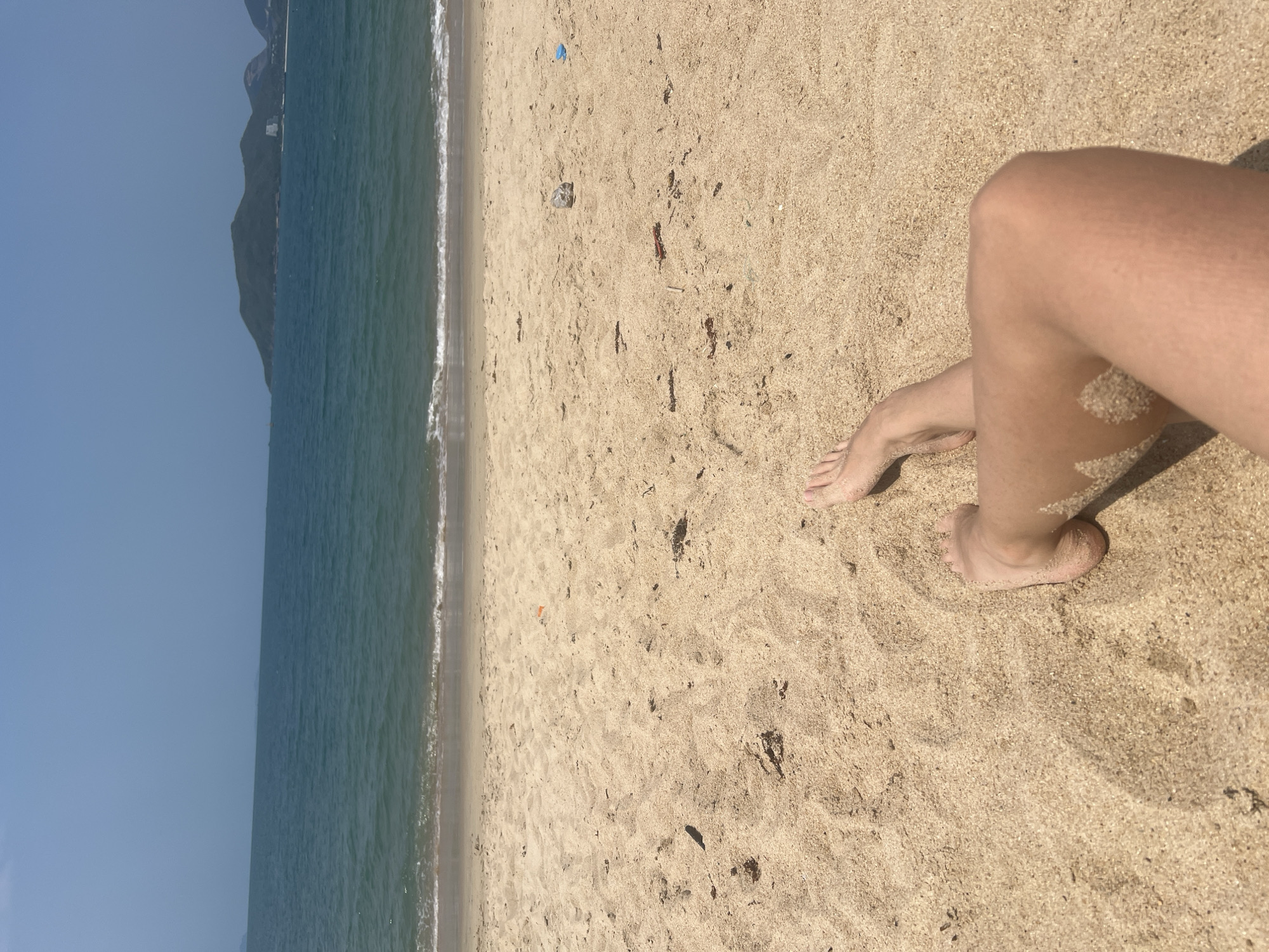 The image captures a coastal scene, possibly in Nha Trang Ward, Vietnam, judging from the visible landmass silhouette. The composition is dominated by the beach, with a wide expanse of sand stretching from the lower right corner towards the center of the frame. The ocean occupies the left side, with a clear horizon line and gentle waves meeting the shore. Human legs and feet are visible in the lower right quadrant, suggesting a person relaxing on the beach. The sand has a light beige color, while the ocean exhibits a blue-green hue.