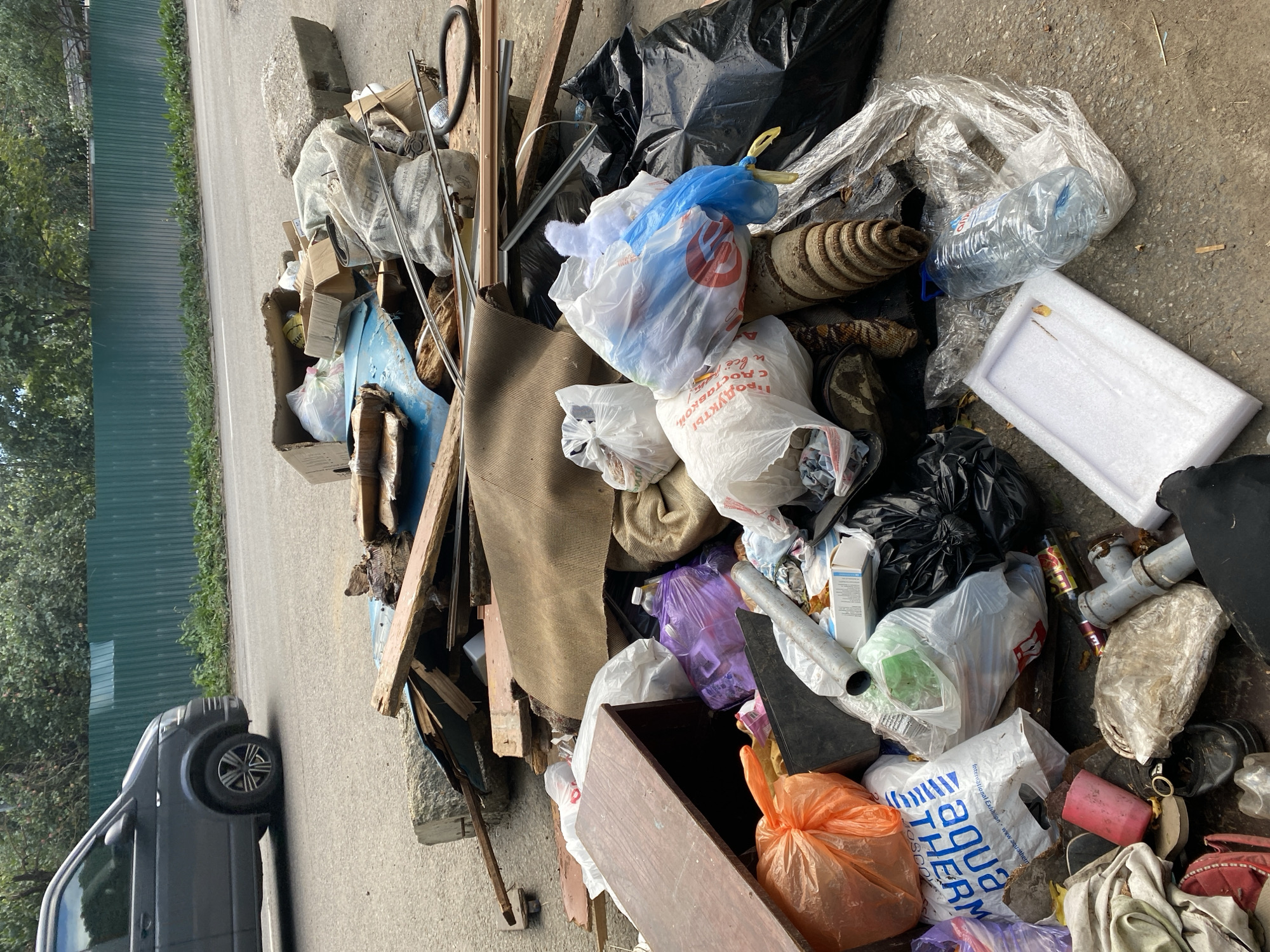 An outdoor, low-angle medium shot depicts a large pile of trash on a paved surface adjacent to a road. The trash consists of various items, including broken wooden planks, cardboard boxes, plastic bags filled with debris, a large plastic water bottle, a rolled-up carpet or mat, and a piece of white styrofoam. Among the refuse are also plumbing pipes and a dark-colored piece of furniture with a wooden surface. In the background, a dark gray van is partially visible driving on the road, and a tall, green metal fence borders the area. The environment appears to be an urban setting, possibly a street or alleyway. The lighting suggests it is daytime.