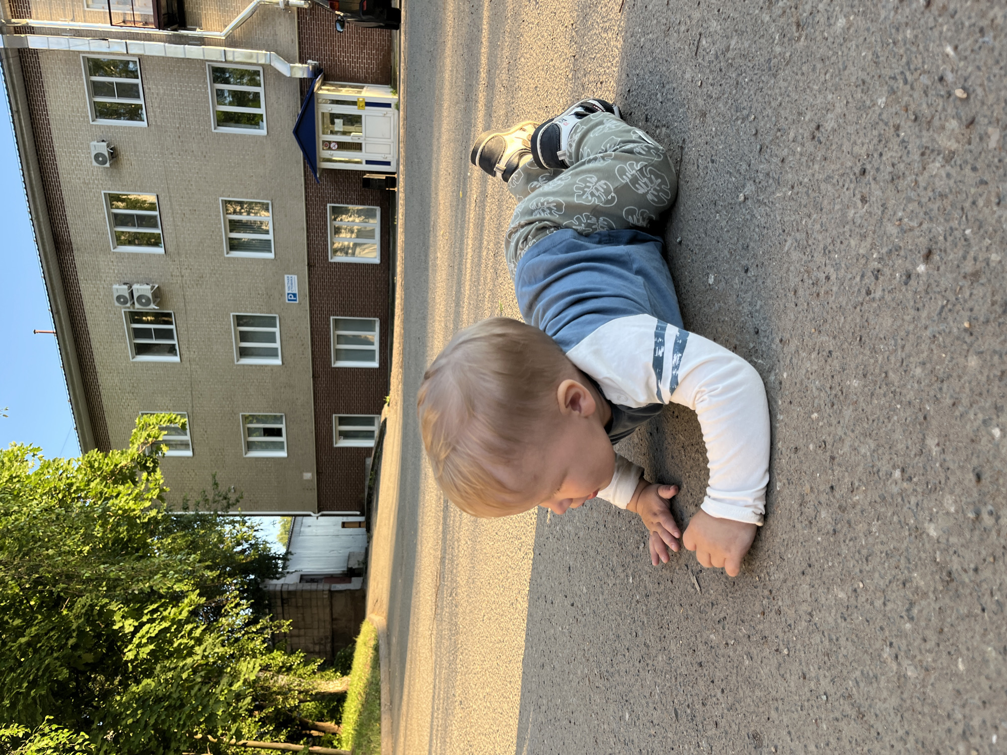 A young Caucasian child, appearing to be approximately 1-2 years old, is lying on its stomach on an asphalt surface. The child has blonde hair and is wearing a long-sleeved light blue shirt with white stripes on the sleeves and light green pants with a leaf pattern. The child's face is turned to the left, and its hands are extended forward on the ground. In the background, a light-colored, multi-story building with several windows and air conditioning units is visible. To the left of the building, there are trees with green foliage and a corrugated metal structure. The scene appears to be outdoors, with sunlight casting shadows on the ground, suggesting it is either early morning or late afternoon. The location is identified as Chaikovsky, Russia.