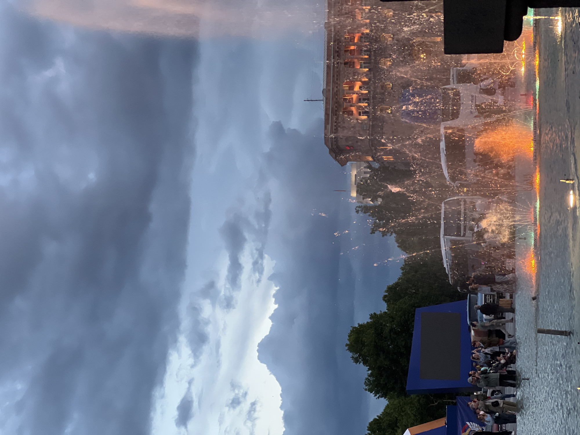 The image captures a public gathering in Yerevan, Armenia, likely in Republic Square, as suggested by the presence of illuminated fountains and a building resembling Government House. The scene is illuminated with decorative lights, with people watching a show with colored lights in the fountain, and some stand near a blue structure, possibly a stage or screen. Dark, dramatic clouds fill the sky, suggesting an evening setting. The water feature is the focal point, illuminated by colored lights. The people are standing, observing, and seem engaged in the spectacle.